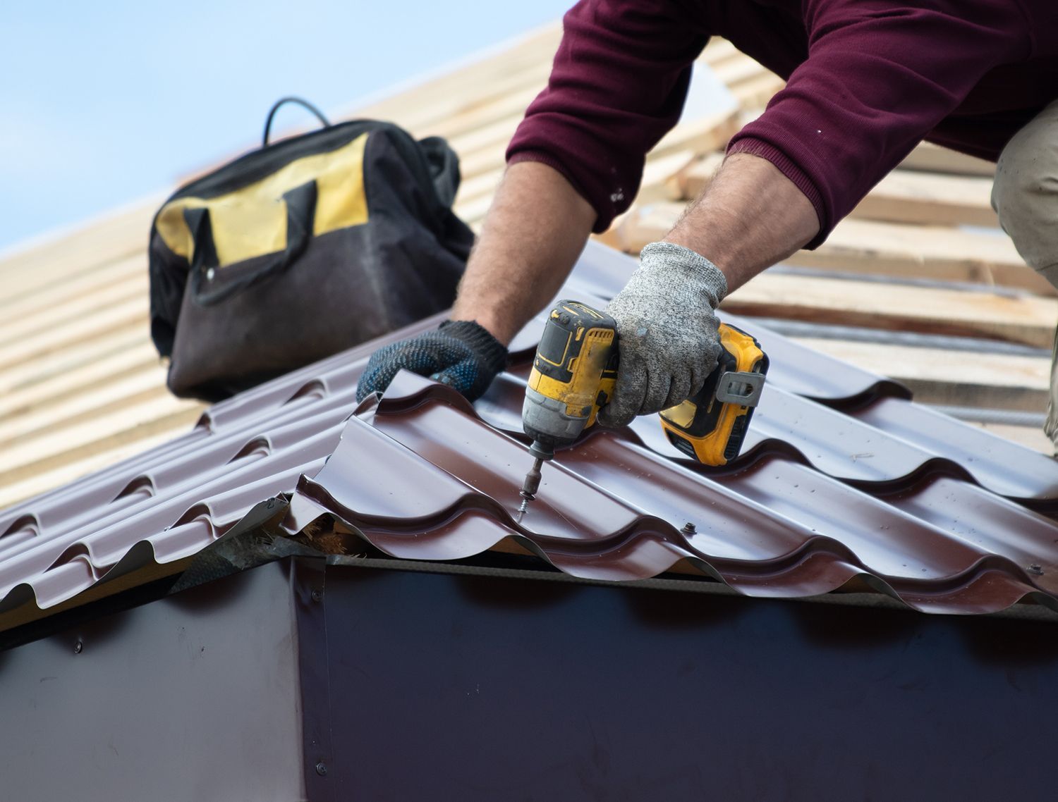 Worker installing brown metal roofing on a roof, using yellow gloves and tools. Worker installing brown metal roofing on a roof, using yellow gloves and tools.