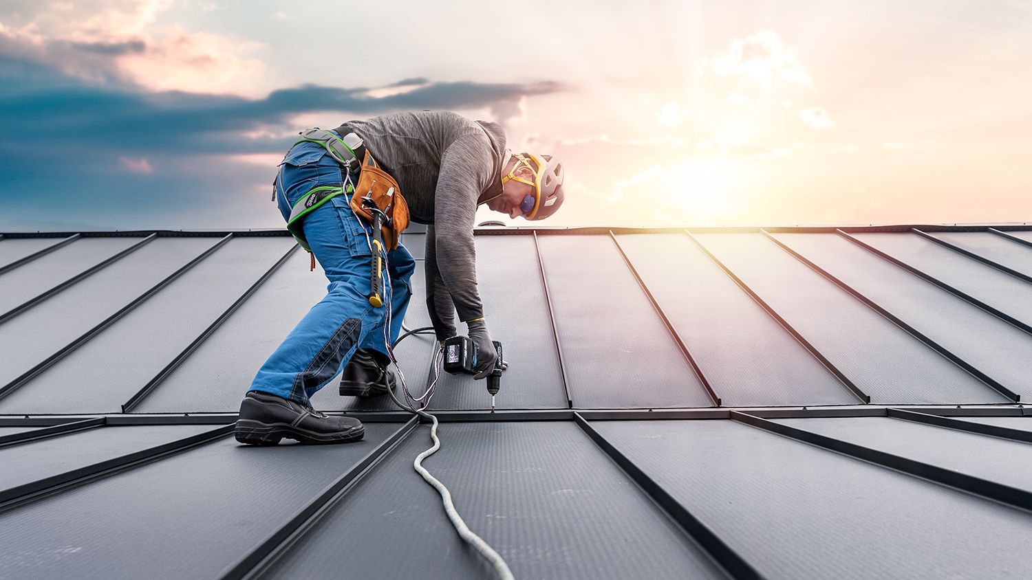 Two roofers installing panels on a metal roof at sunrise, with tools and safety gear visible. Two roofers installing panels on a metal roof at sunrise, with tools and safety gear visible.