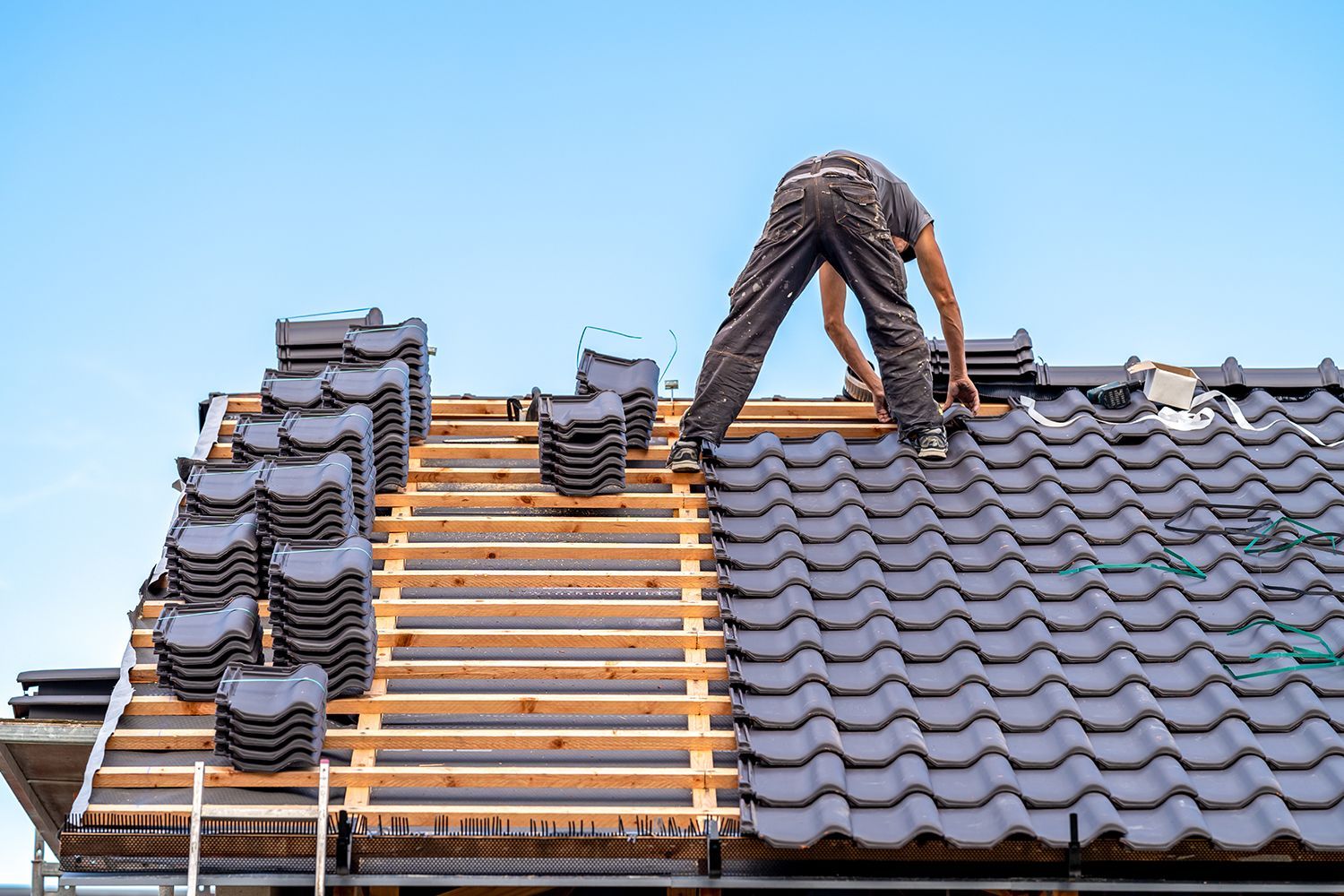 Worker installing roof tiles on a house roof with stacked tiles and exposed wooden battens Worker installing roof tiles on a house roof with stacked tiles and exposed wooden battens