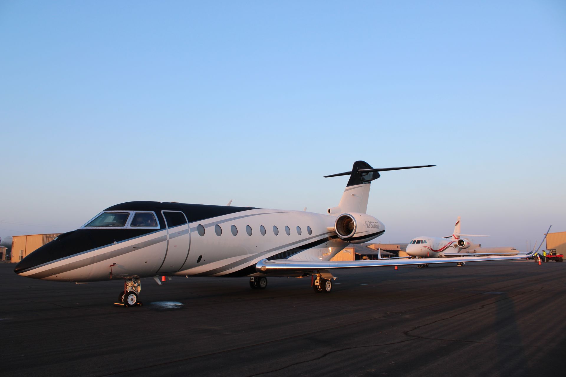 A white and black private jet parked on an airport tarmac under a clear sky, with another aircraft visible behind it.