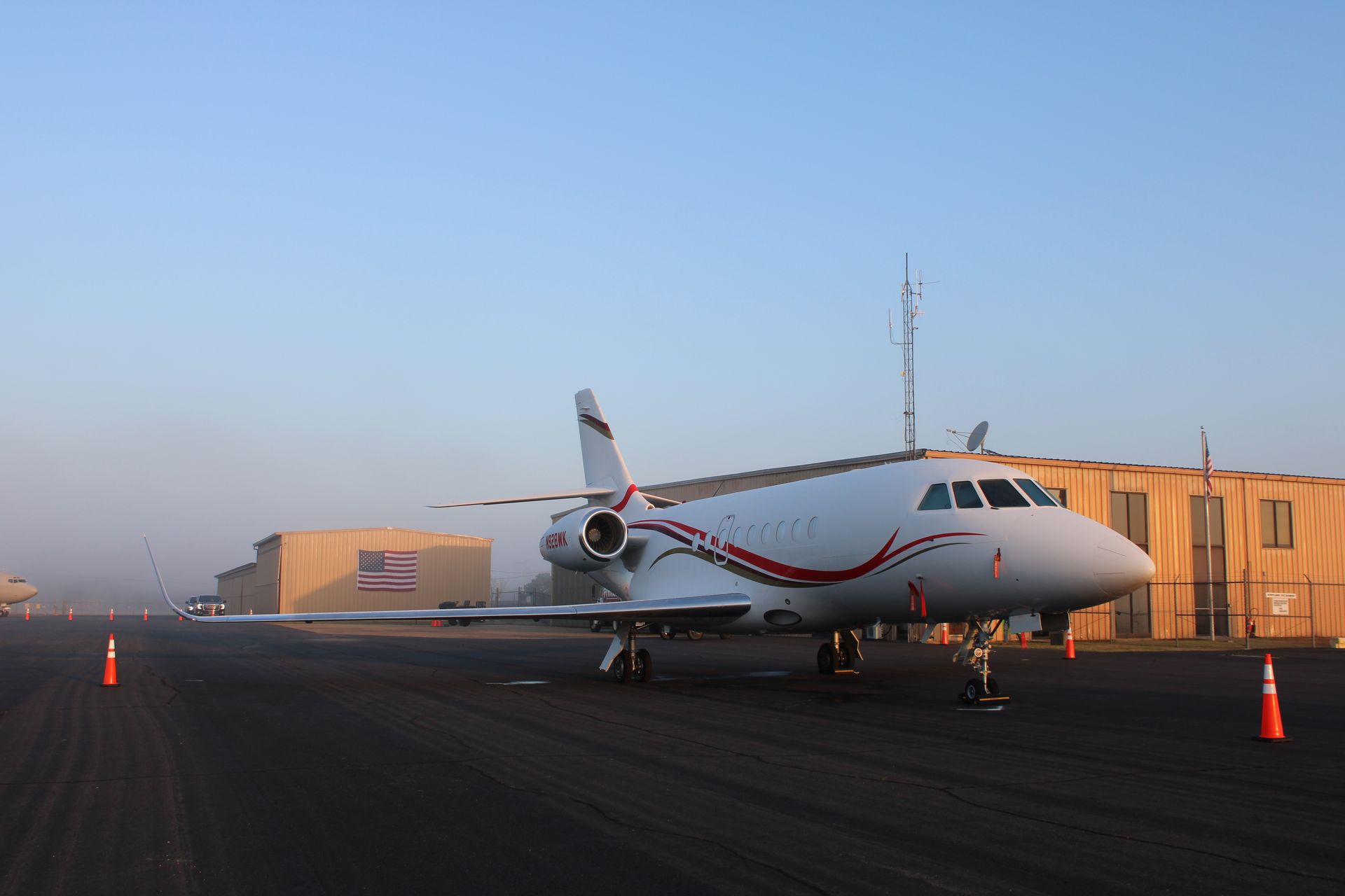 A white private jet with red accents parked on an airport tarmac near buildings at dawn.