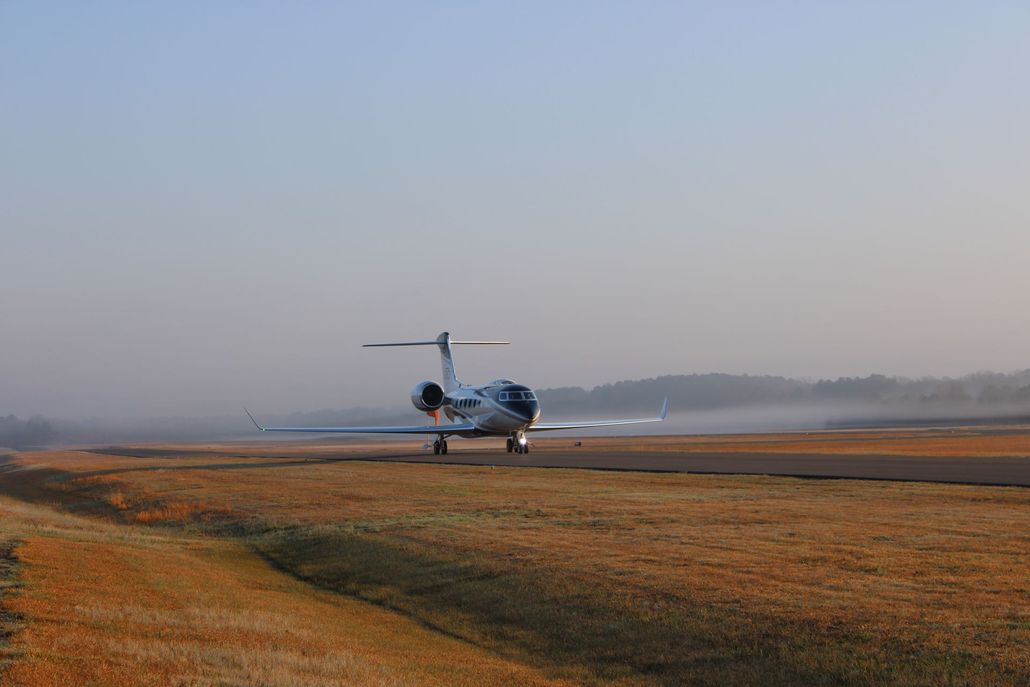 A private business jet taxis on an airport runway surrounded by dry, golden grass under a hazy, pale sky.