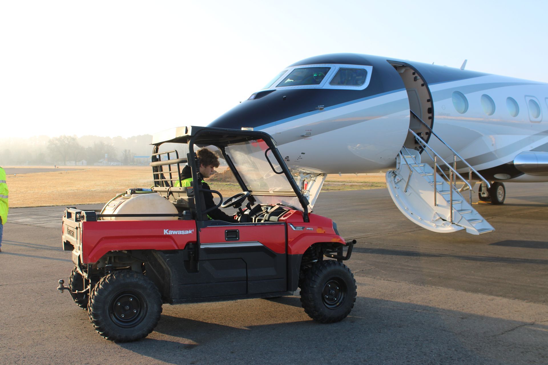 A person drives a red utility vehicle past the open door and steps of a parked private jet on an airport tarmac.
