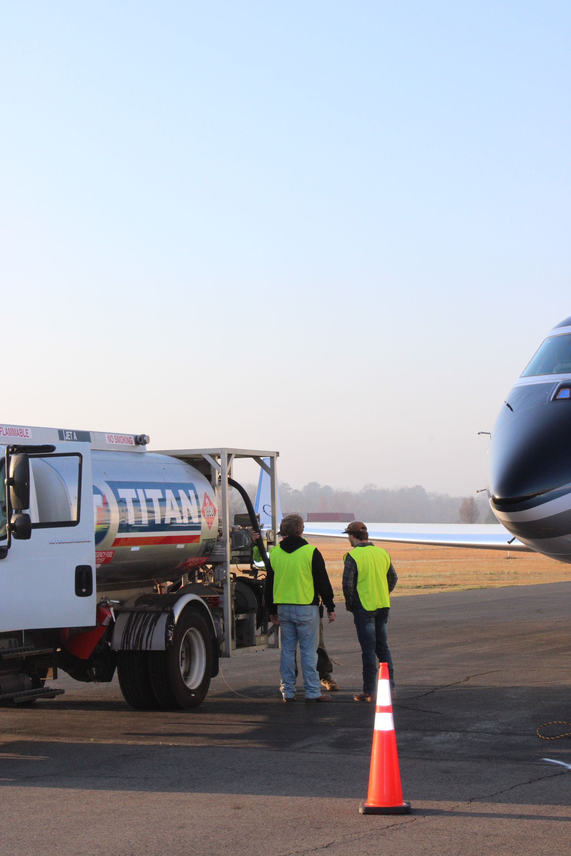 Two people in bright yellow safety vests stand by a Titan aviation fuel truck on an airport tarmac next to a plane.