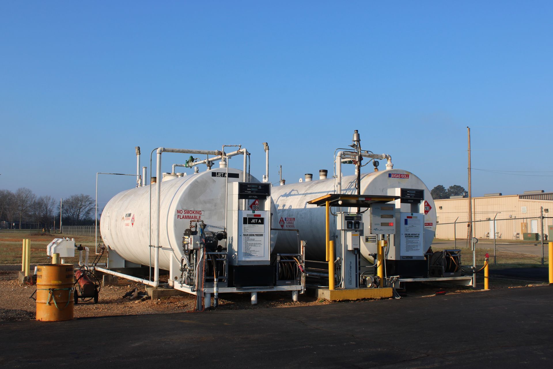 Two large, white cylindrical fuel tanks with attached dispensing pumps sit outdoors against a clear blue sky.