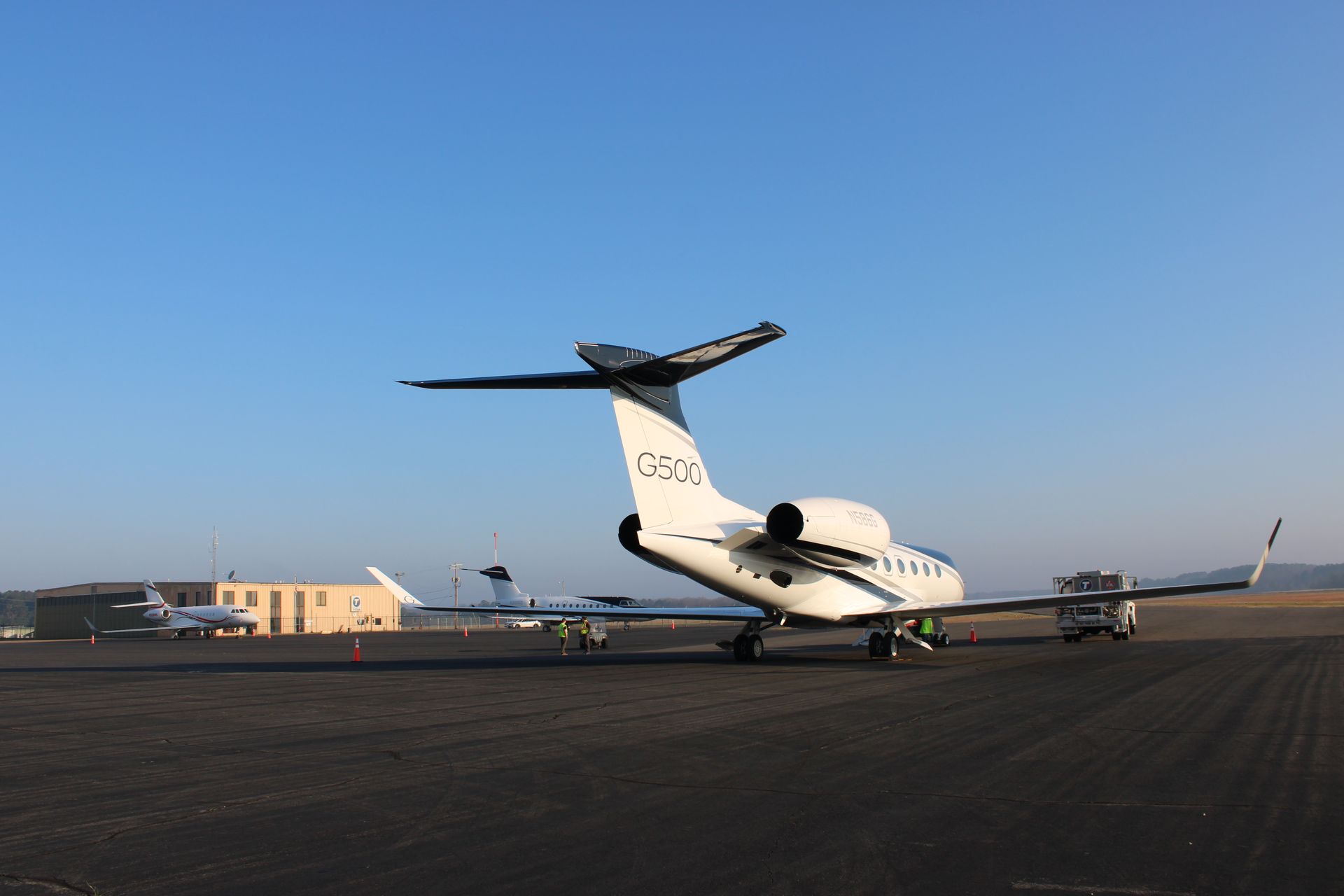A white private jet is parked on an airport tarmac under a clear blue sky, with other aircraft visible in the distance.