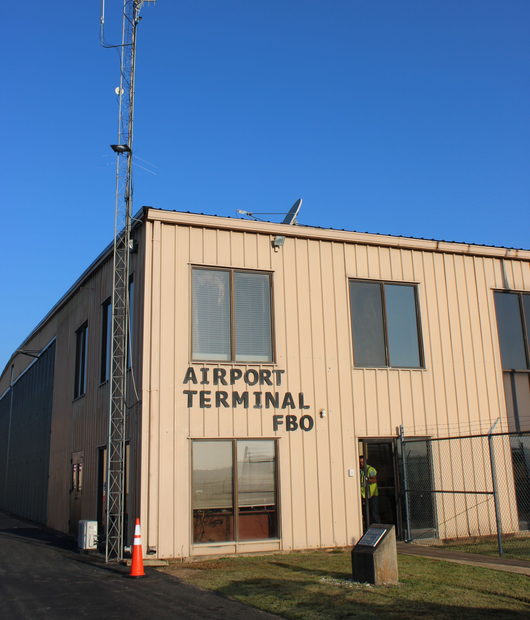 Small beige airport terminal building with “AIRPORT TERMINAL FBO” sign and antennas against a blue sky