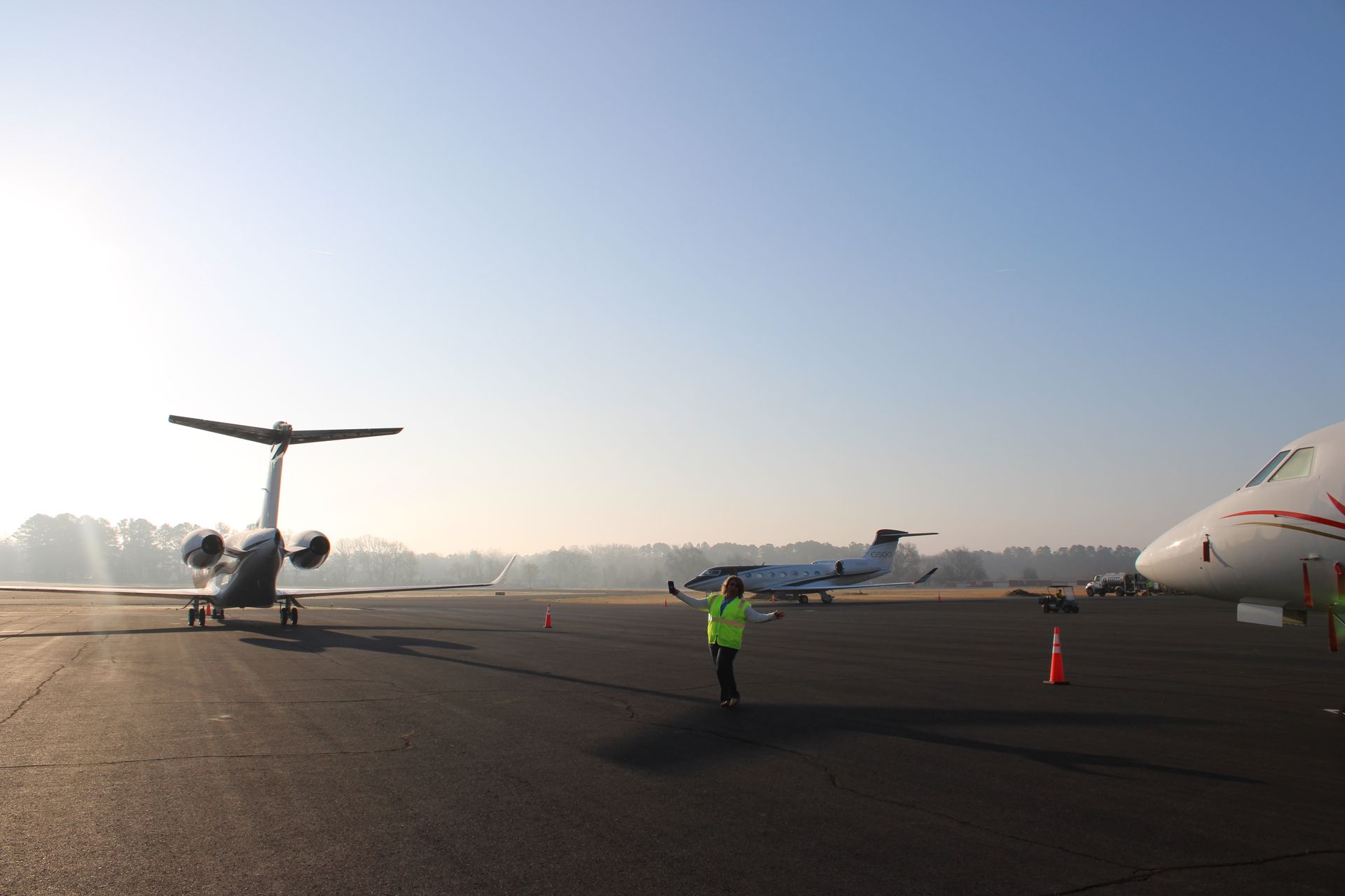 A ground crew member in a bright vest stands on an airport tarmac near two parked private jets on a sunny, hazy day.