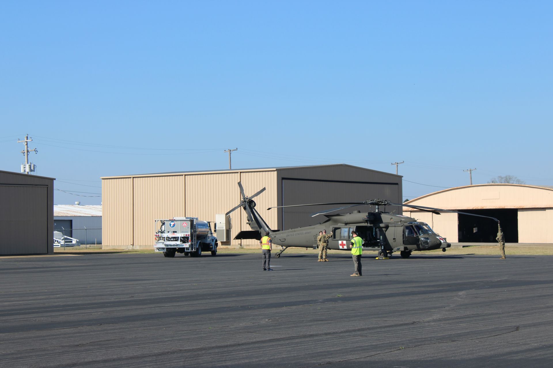 A dark green military helicopter sits on a tarmac near hangars with personnel and a service truck nearby.