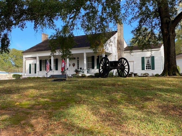 Historic white house with black shutters and large cannon displayed on the lawn, under oak trees.