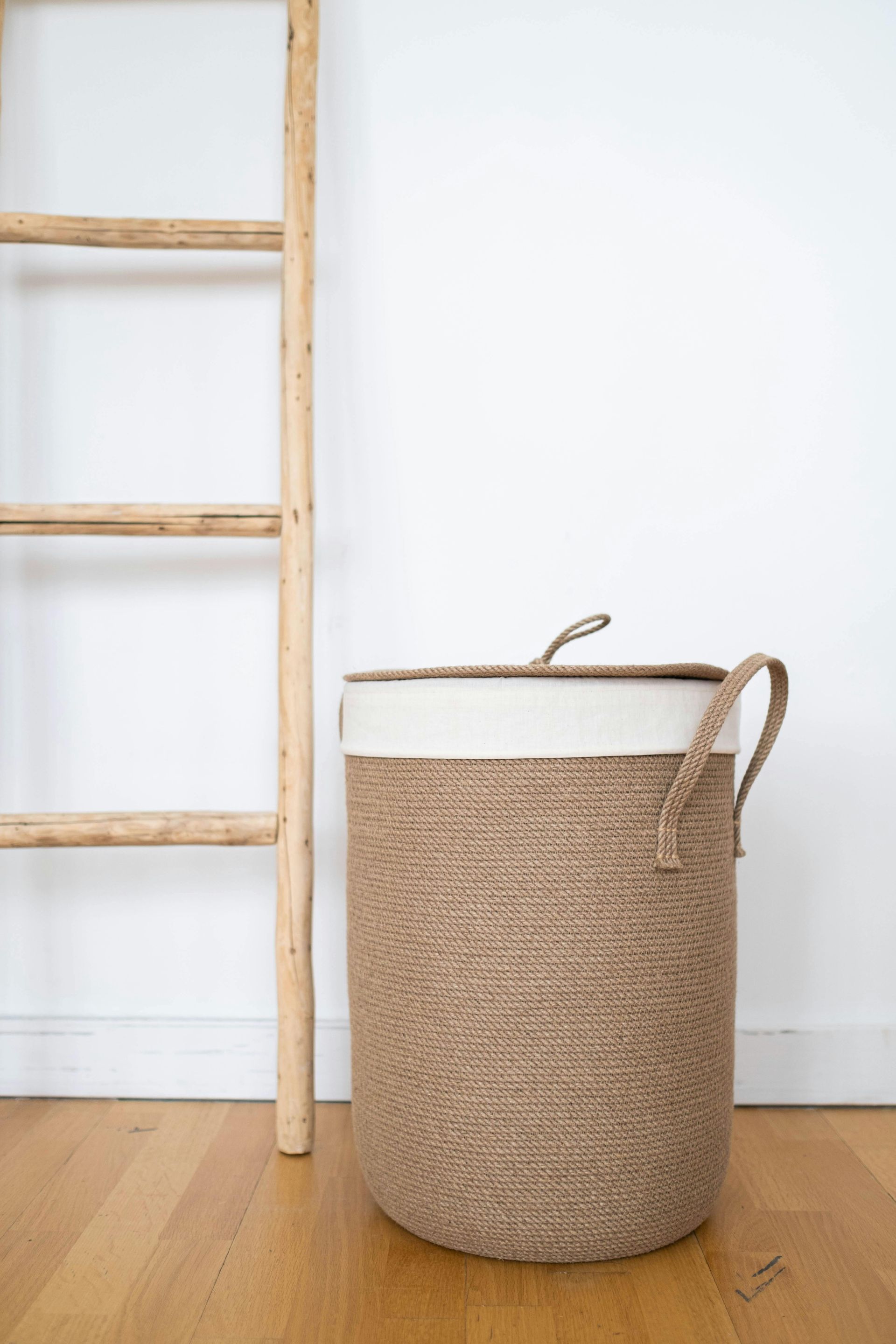 Woven laundry basket with lid and handles, next to a wooden ladder, against a white wall and hardwood floor.