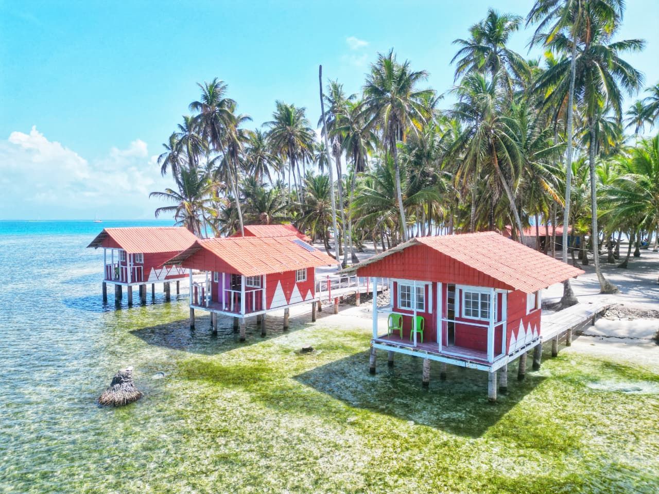 Red beach huts on stilts beside a turquoise sea, with palm trees and a sandy shore in Misdub island
