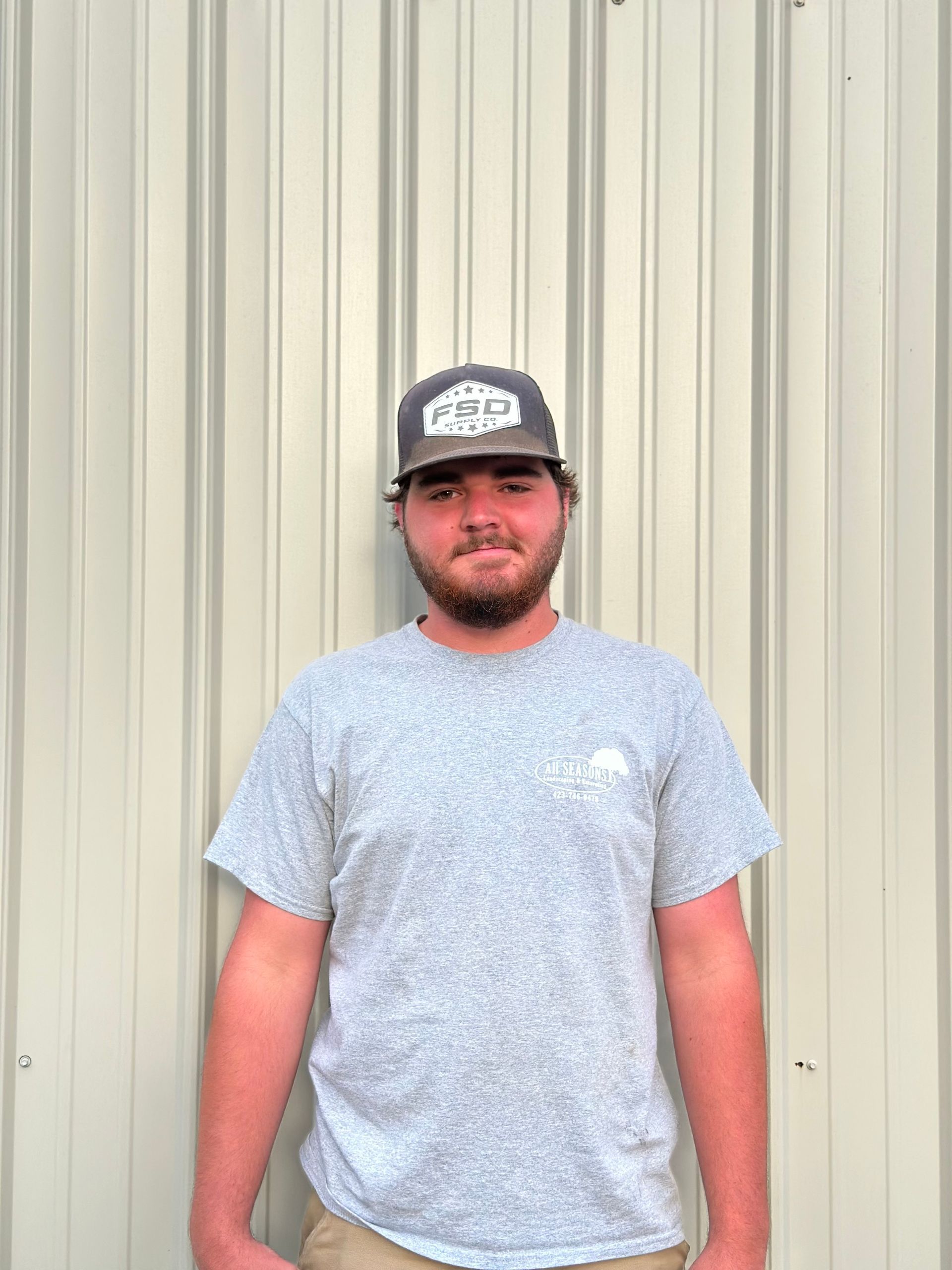 Young man with a beard wearing a hat and gray t-shirt stands in front of a corrugated metal wall.