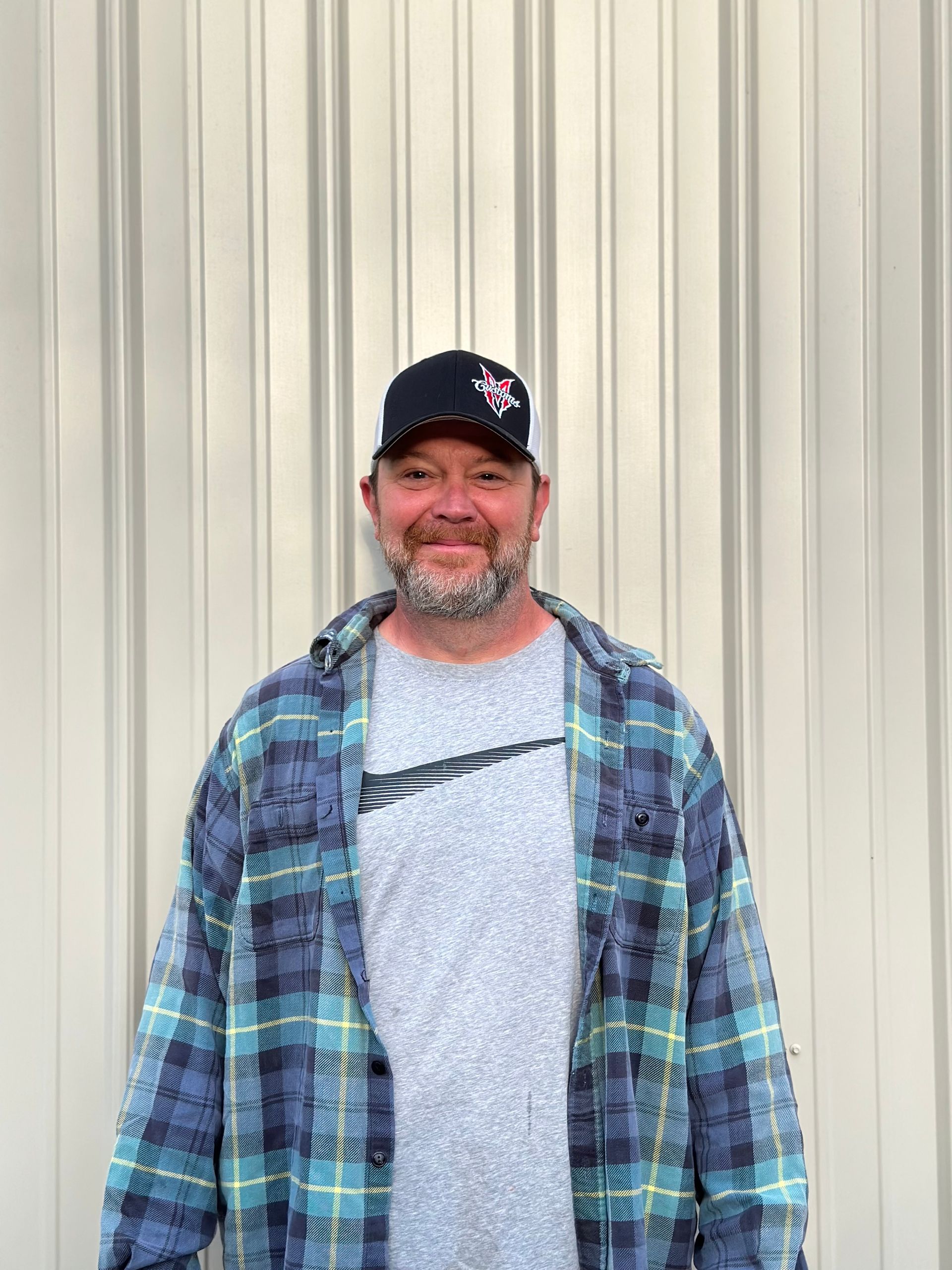Man wearing a cap, plaid shirt, and grey t-shirt, standing in front of a corrugated metal wall.