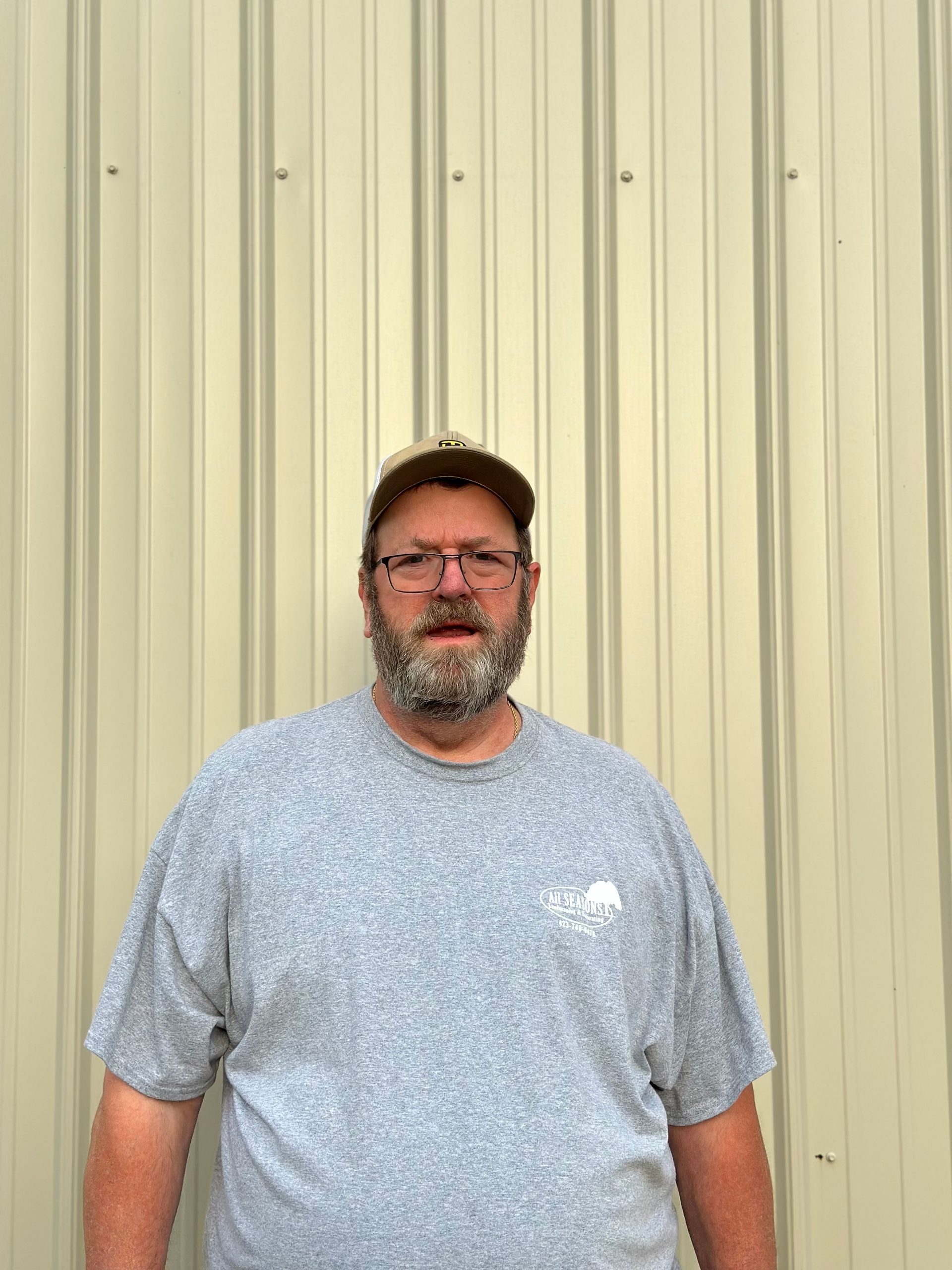 Man with beard and glasses in a gray t-shirt and cap, standing in front of a corrugated metal wall.