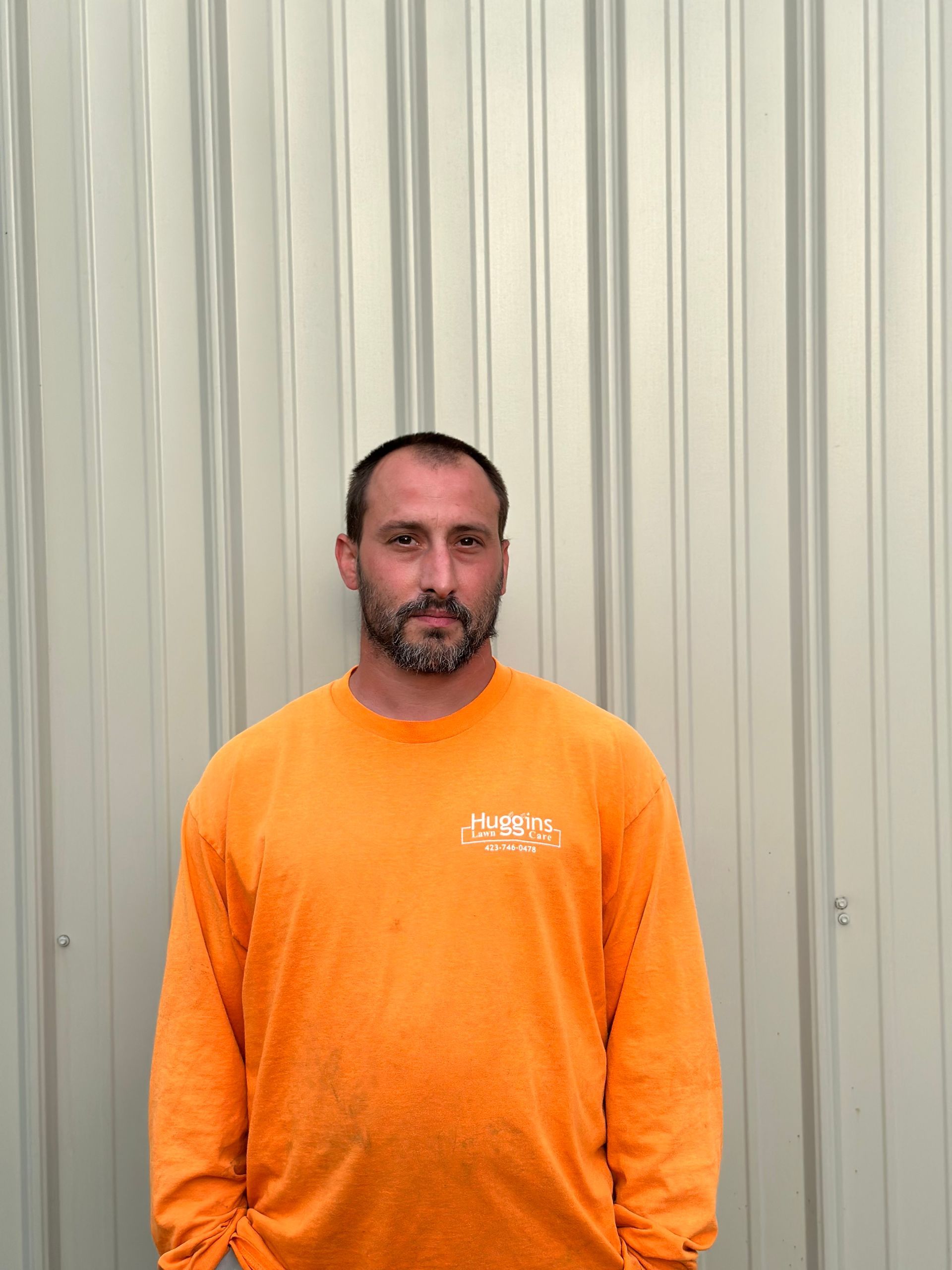 Man in orange long-sleeve shirt stands in front of a corrugated metal wall.