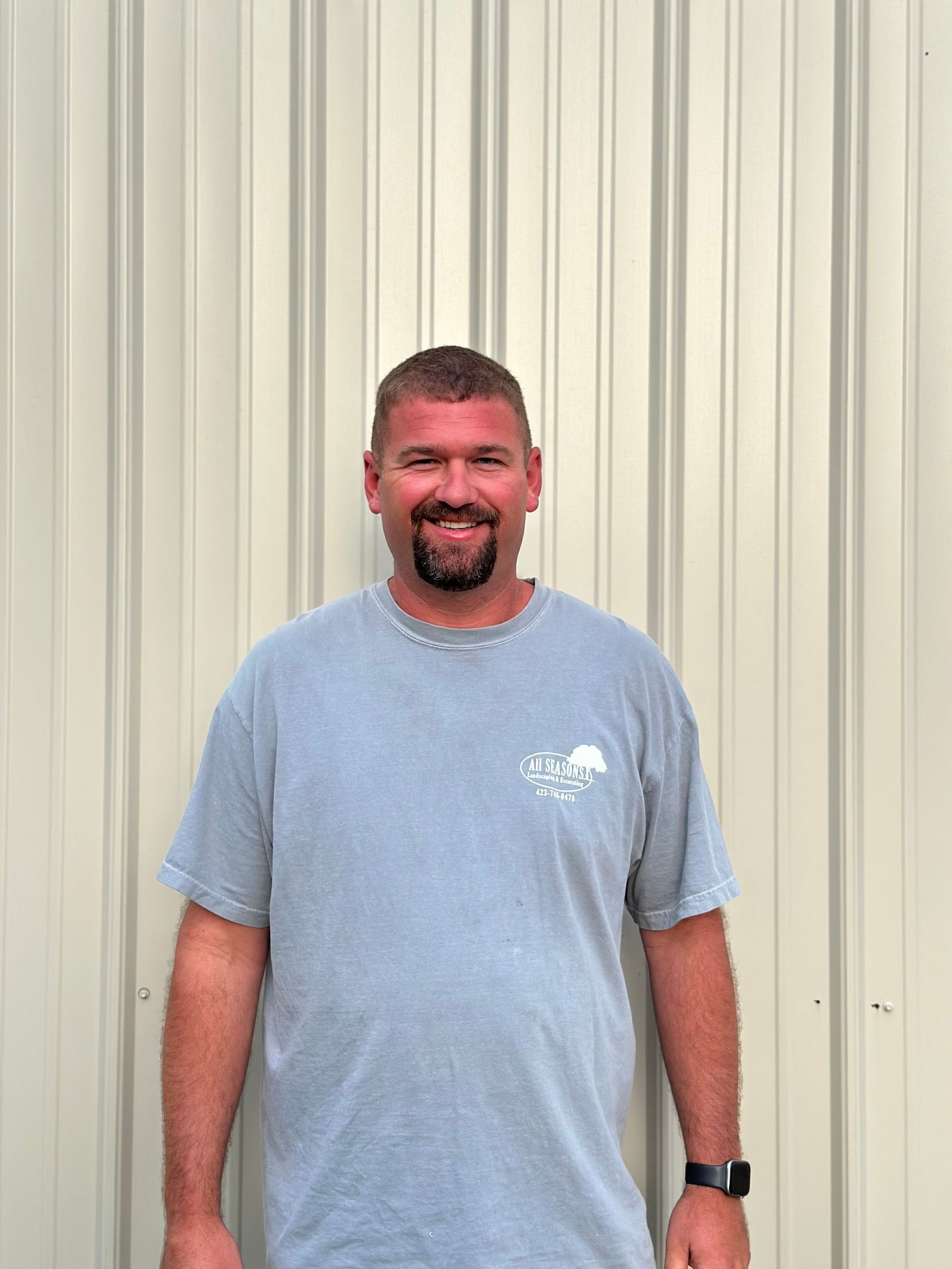 Man with short dark hair and beard smiling, wearing a gray t-shirt, standing in front of a beige wall.
