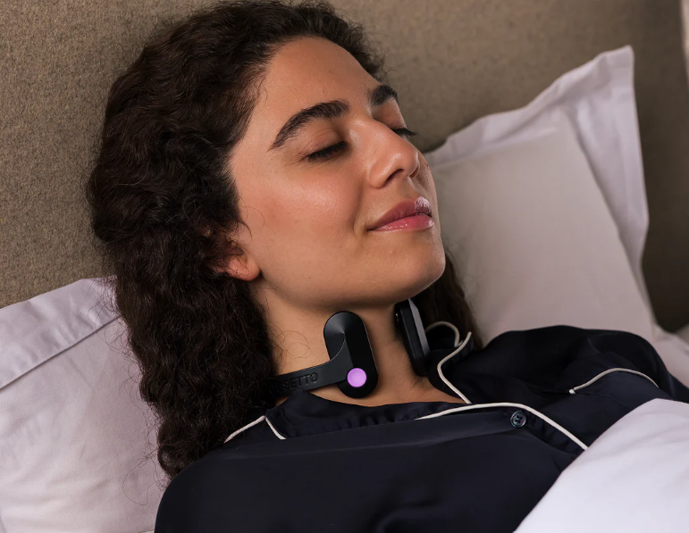 Relaxed person lying in bed with a neck massager, eyes closed, wearing dark pajamas.