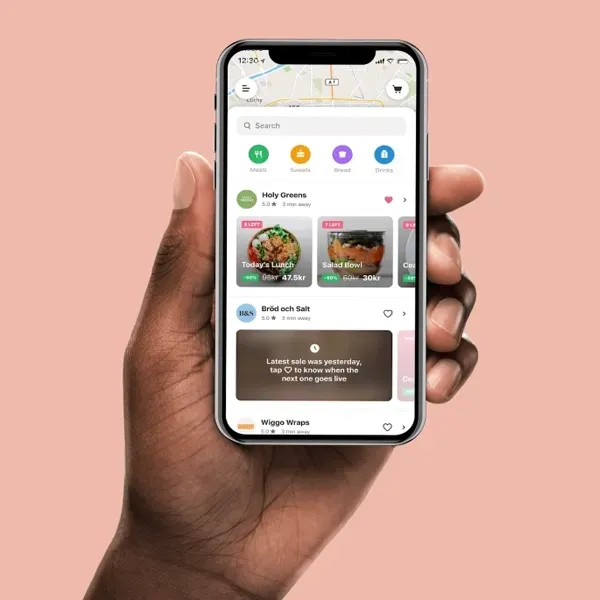A person’s hand holds a smartphone displaying a food delivery app interface against a light pink background.