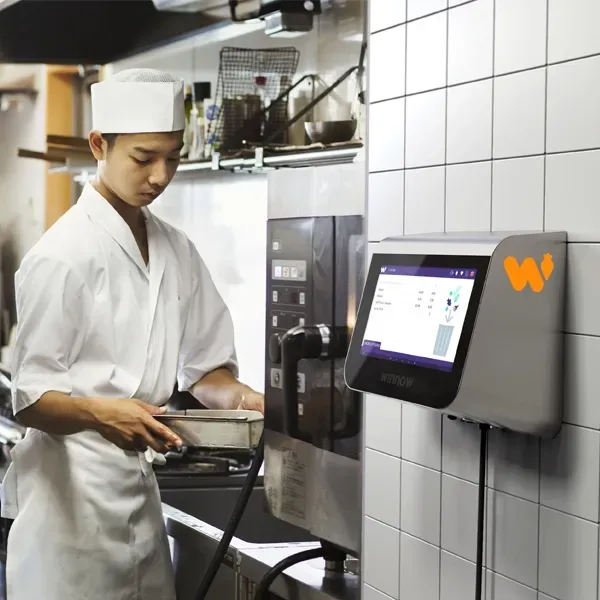A chef works in a kitchen next to a wall-mounted digital monitoring system with an orange logo.