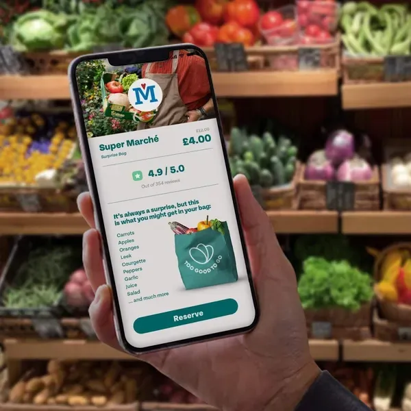 A hand holds a smartphone displaying a food-saving app interface, set against a blurred background of a grocery store.