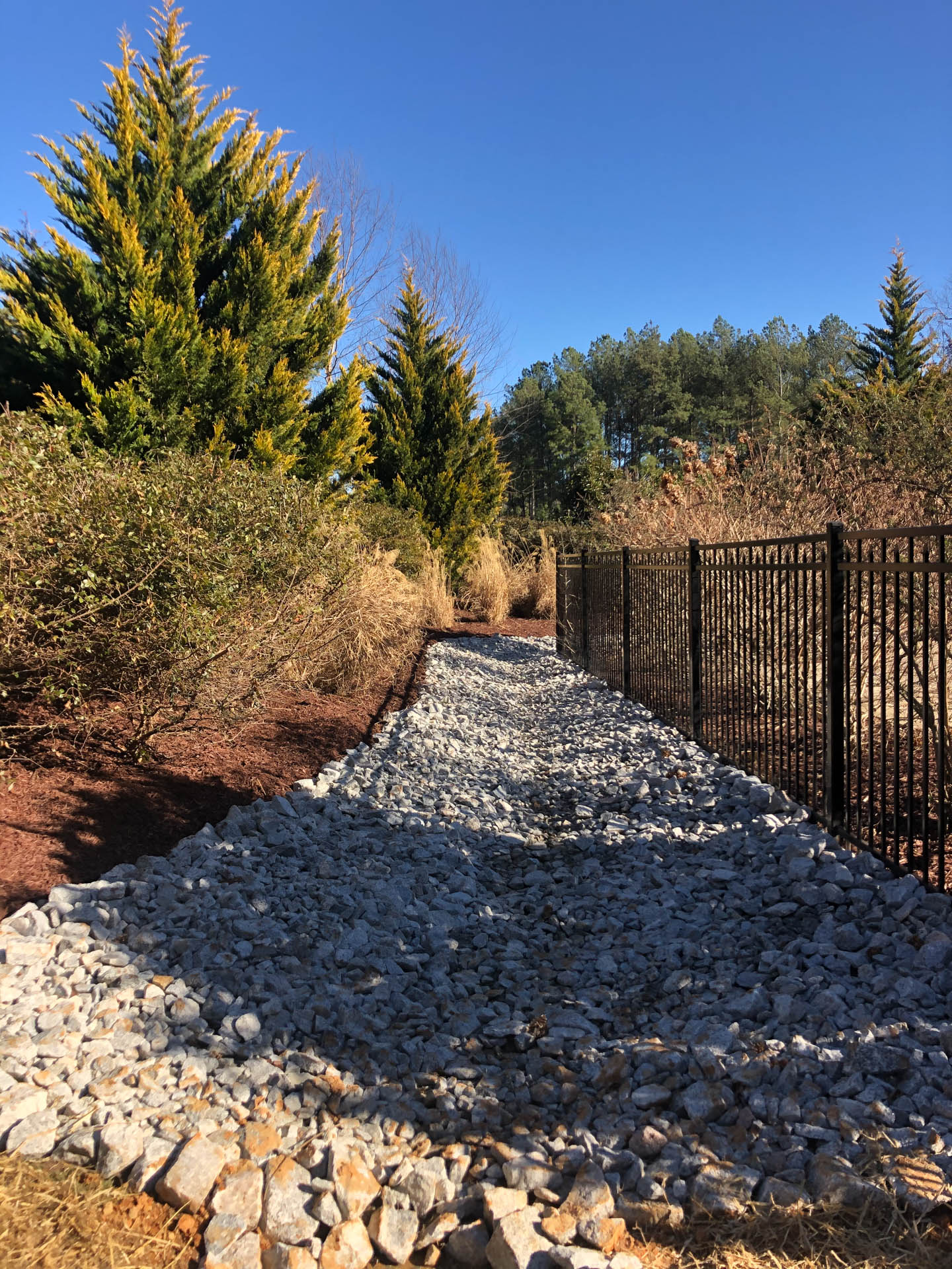 Gravel-lined walkway beside a black fence, with landscaping including trees and bushes under a clear blue sky.