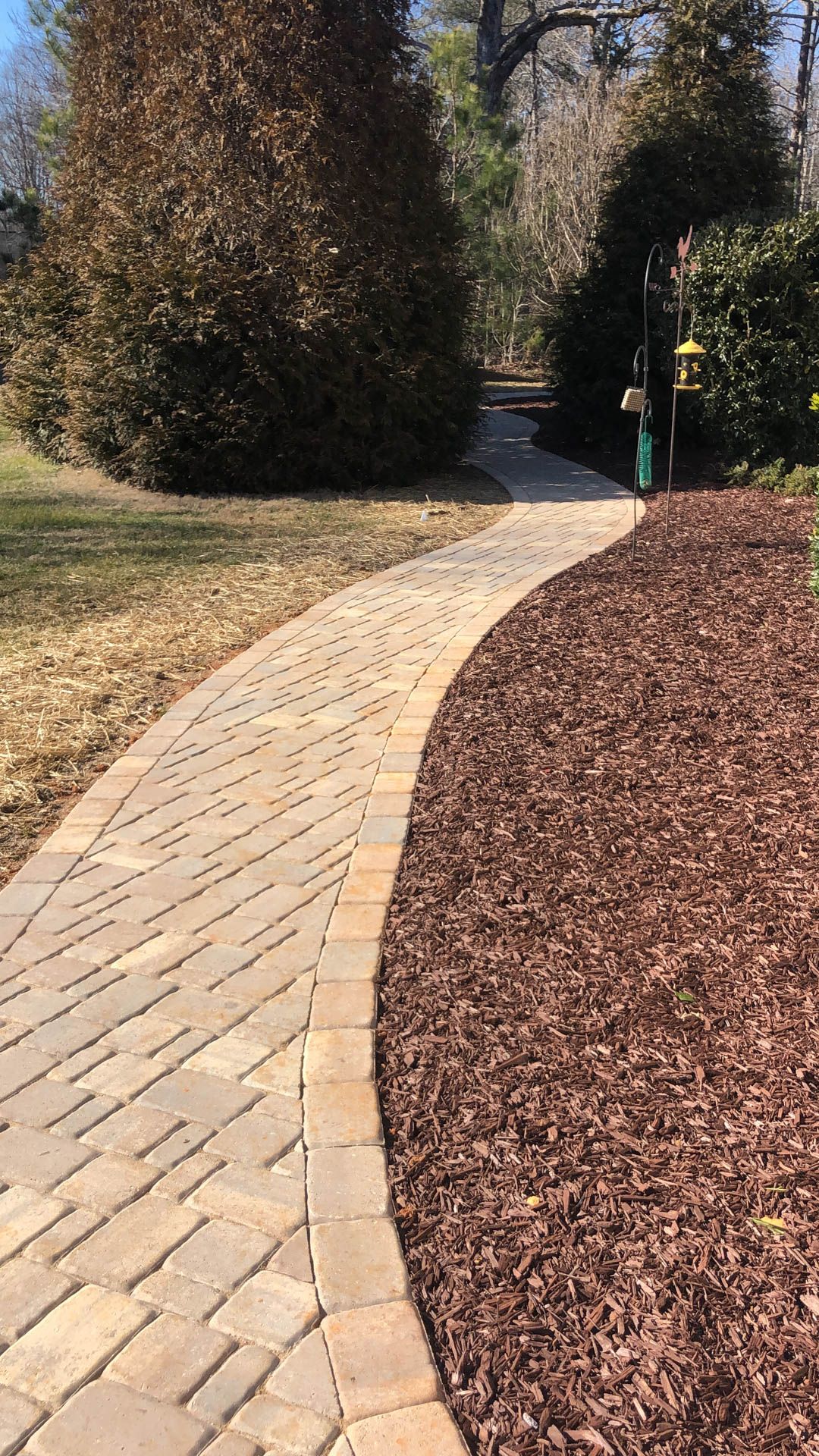 Brick pathway curves through a yard, bordered by dark mulch and a strip of grass. Trees frame the path.