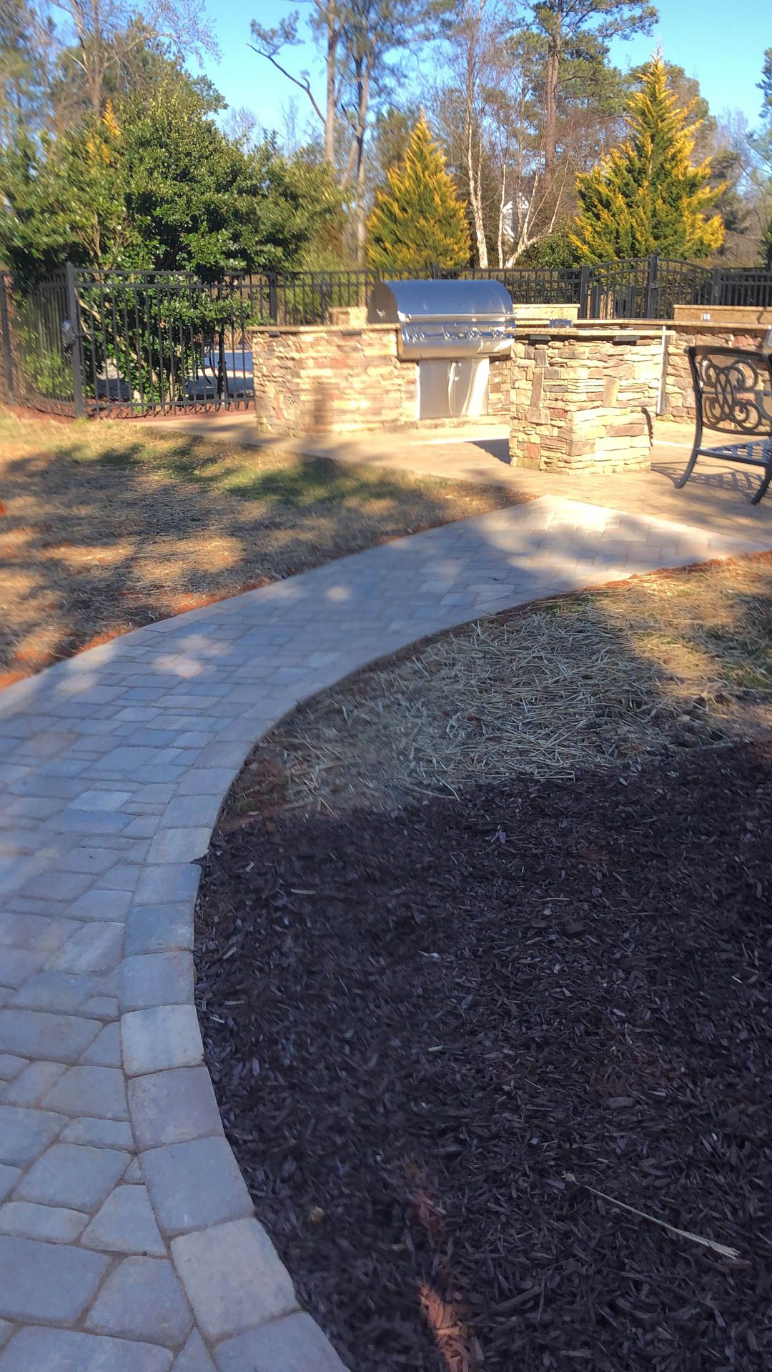 Brick pathway curves towards an outdoor grilling station set into a stone wall.