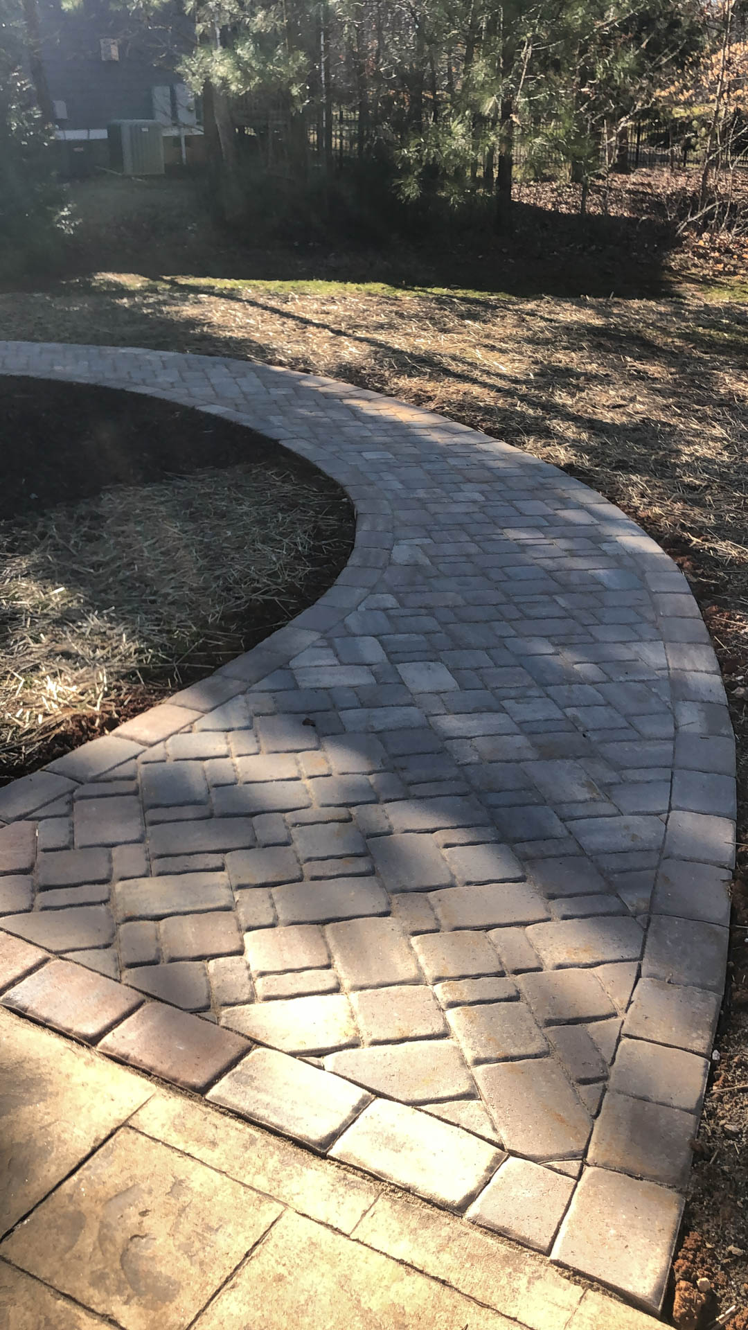 Curved brick paver pathway in a yard, leading toward trees and sunlight.