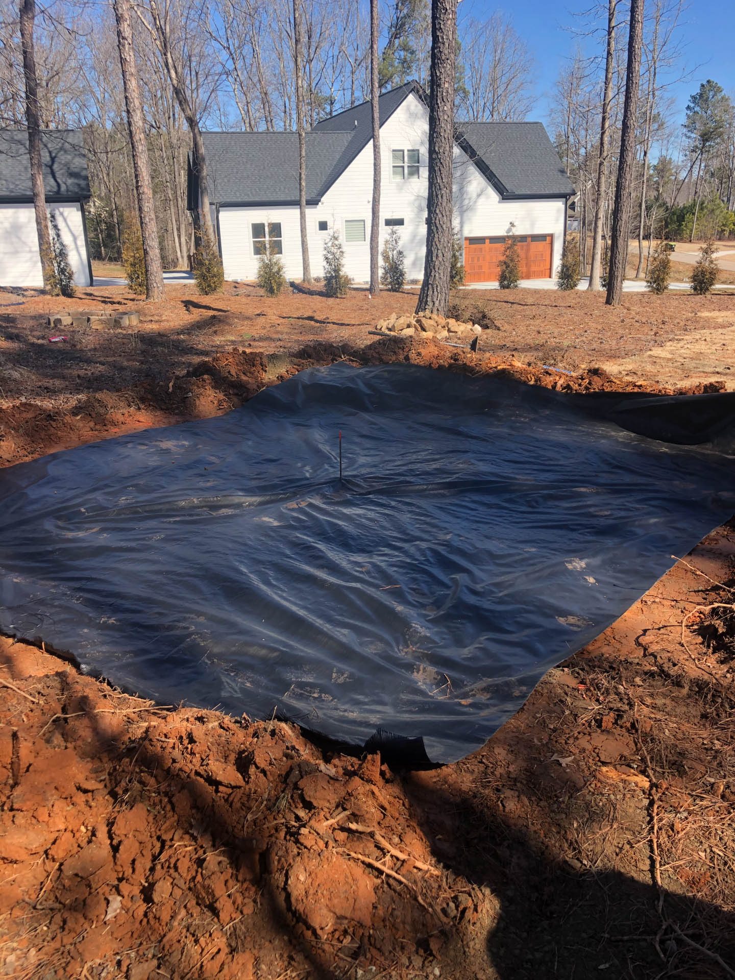 Construction site with black tarp in an excavated pit, with a white house in the background.