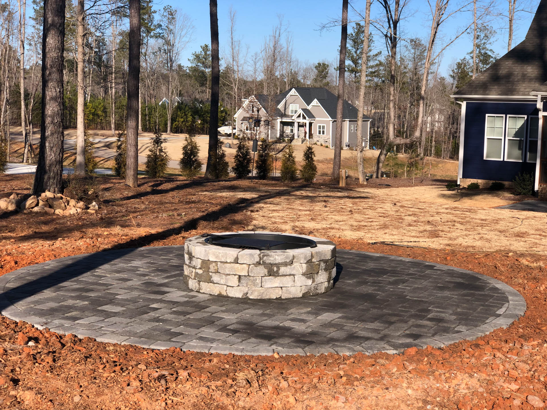 Fire pit on a circular stone patio in a backyard with a house in the background.