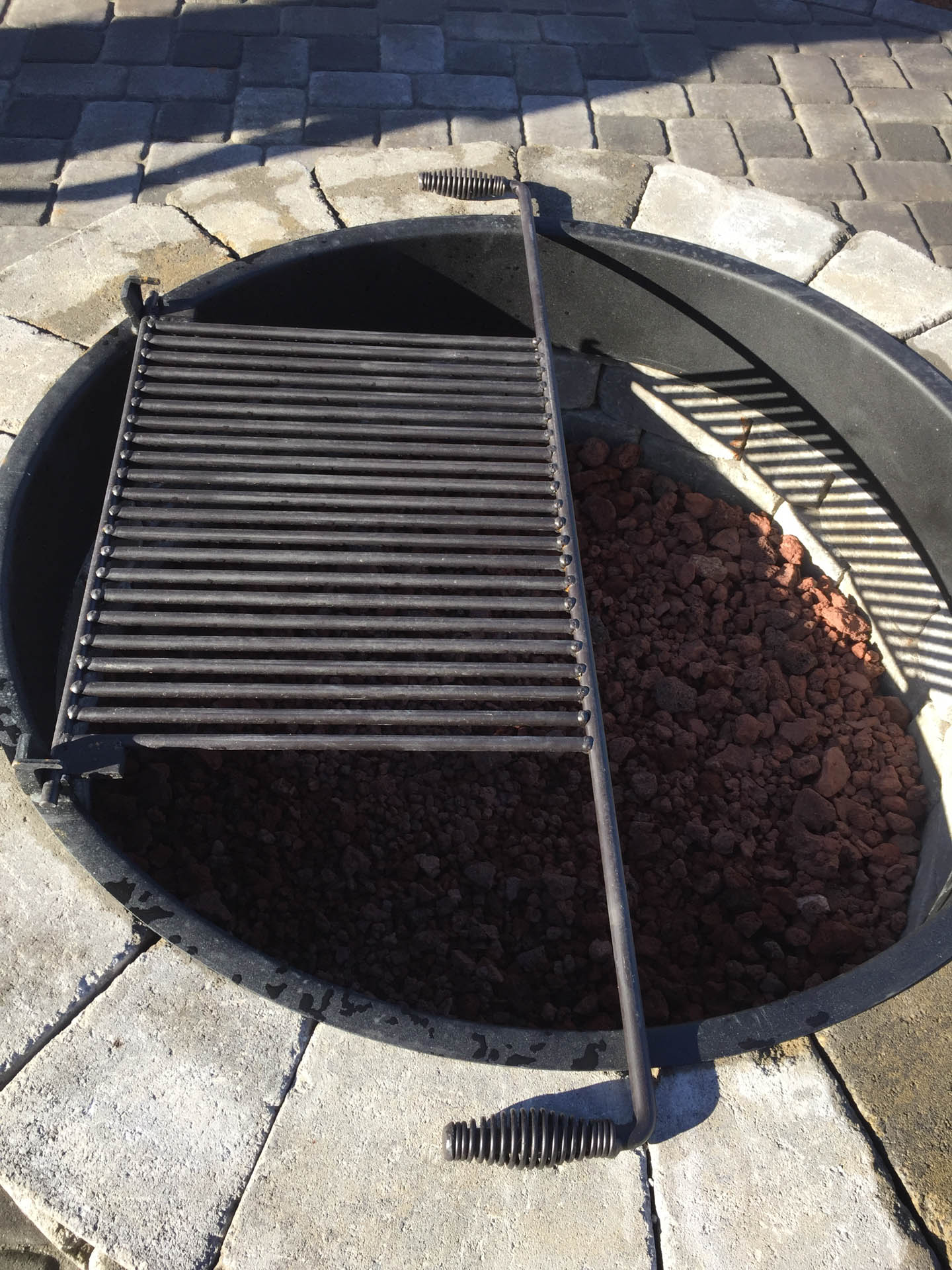 Fire pit with grill. Black metal grill partially covering red lava rocks in a circular pit surrounded by pavers.