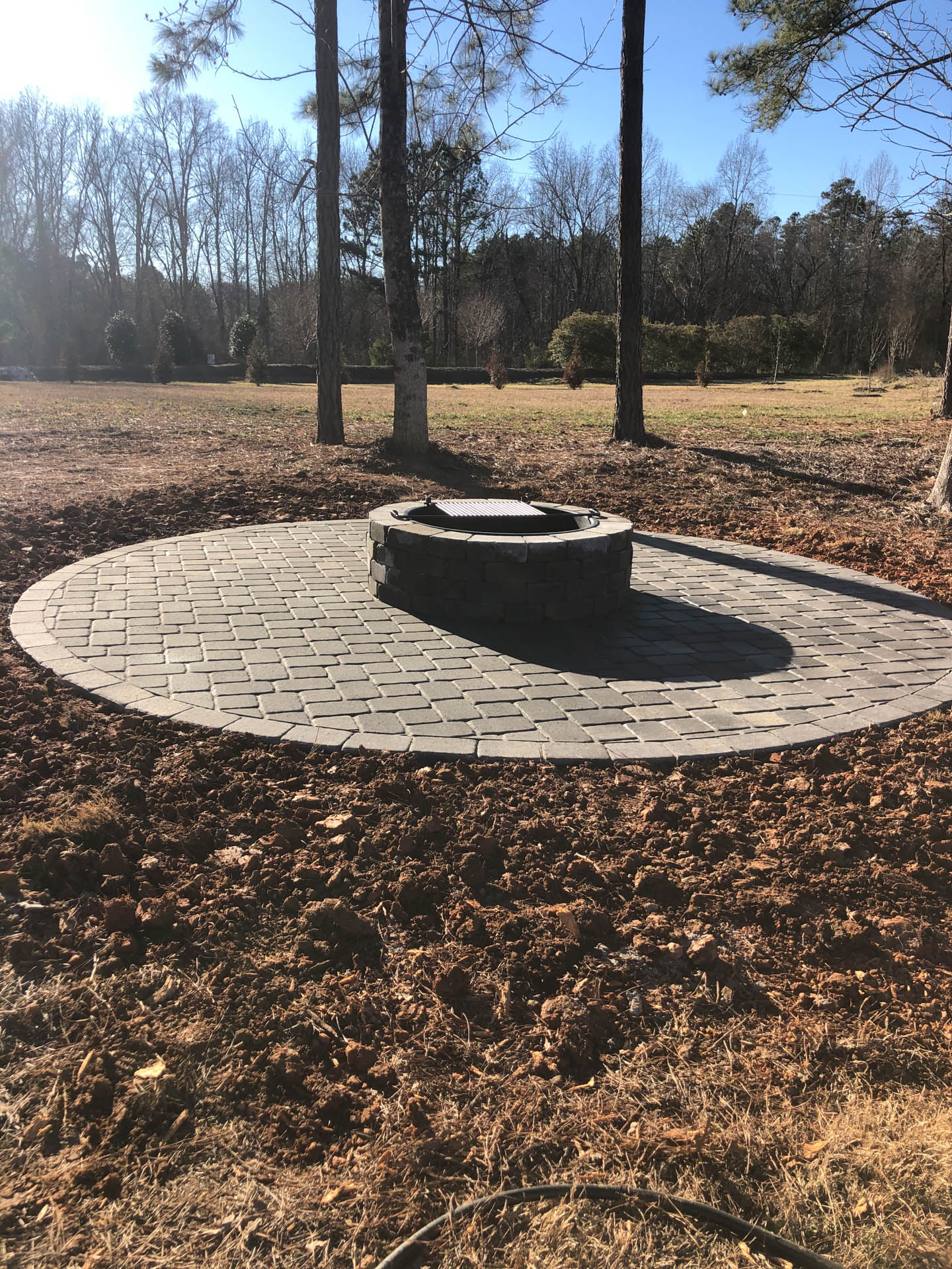 A circular stone patio with a fire pit in a yard, surrounded by brown leaves and trees under a clear sky.