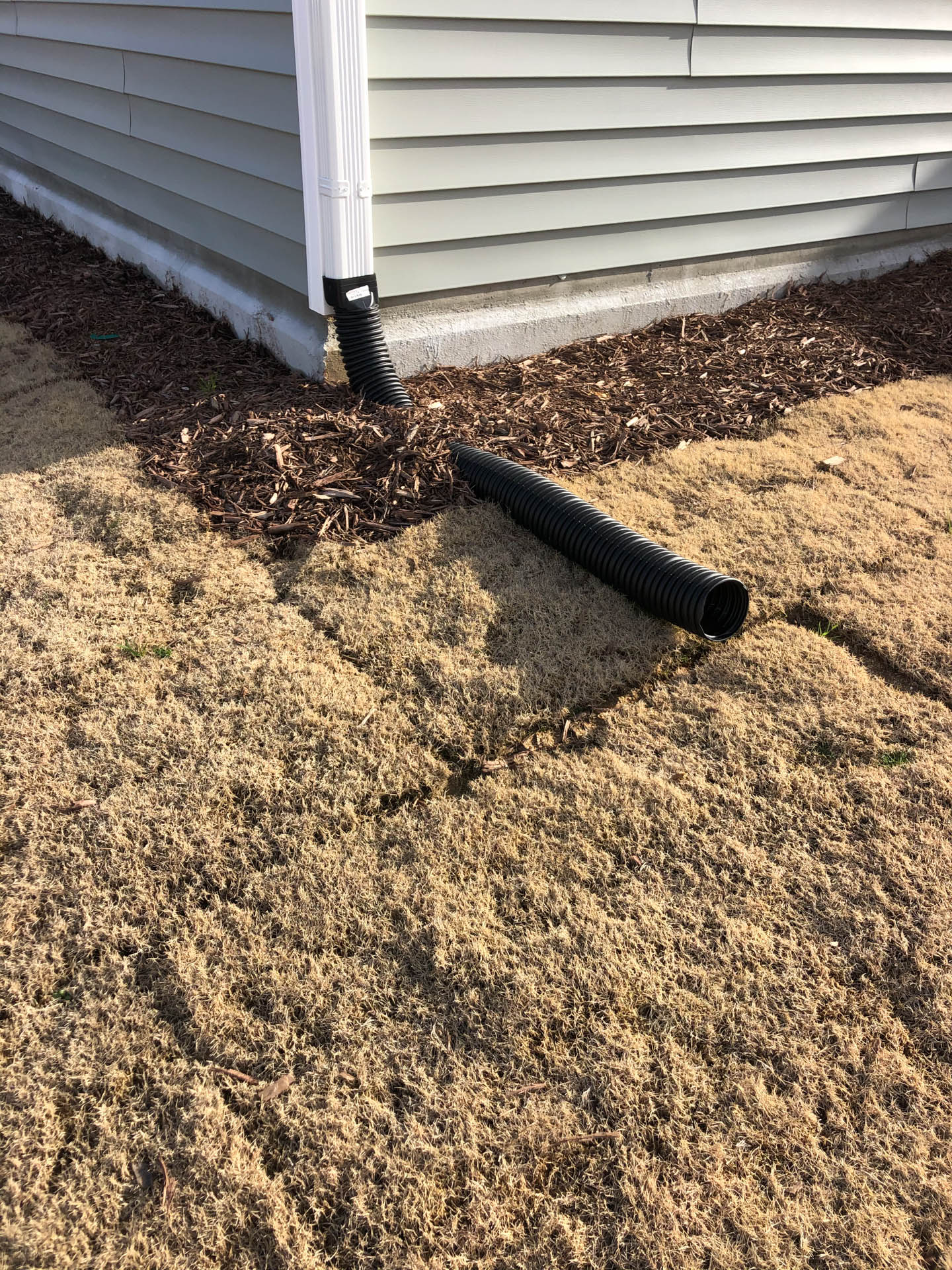 Downspout directing water to a black drainage pipe on a lawn with dry brown grass, near a building.