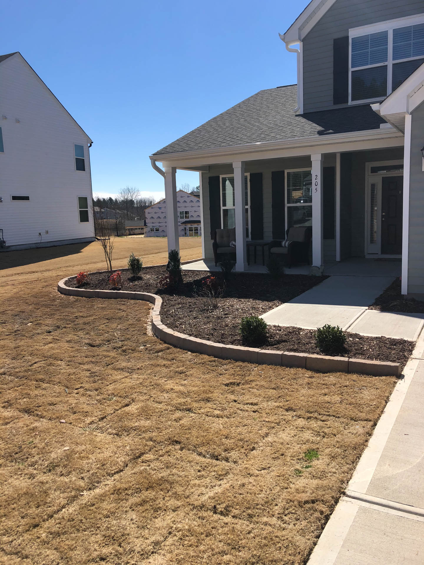 A two-story house with a porch. Brown lawn, dark mulch beds, and blue sky.