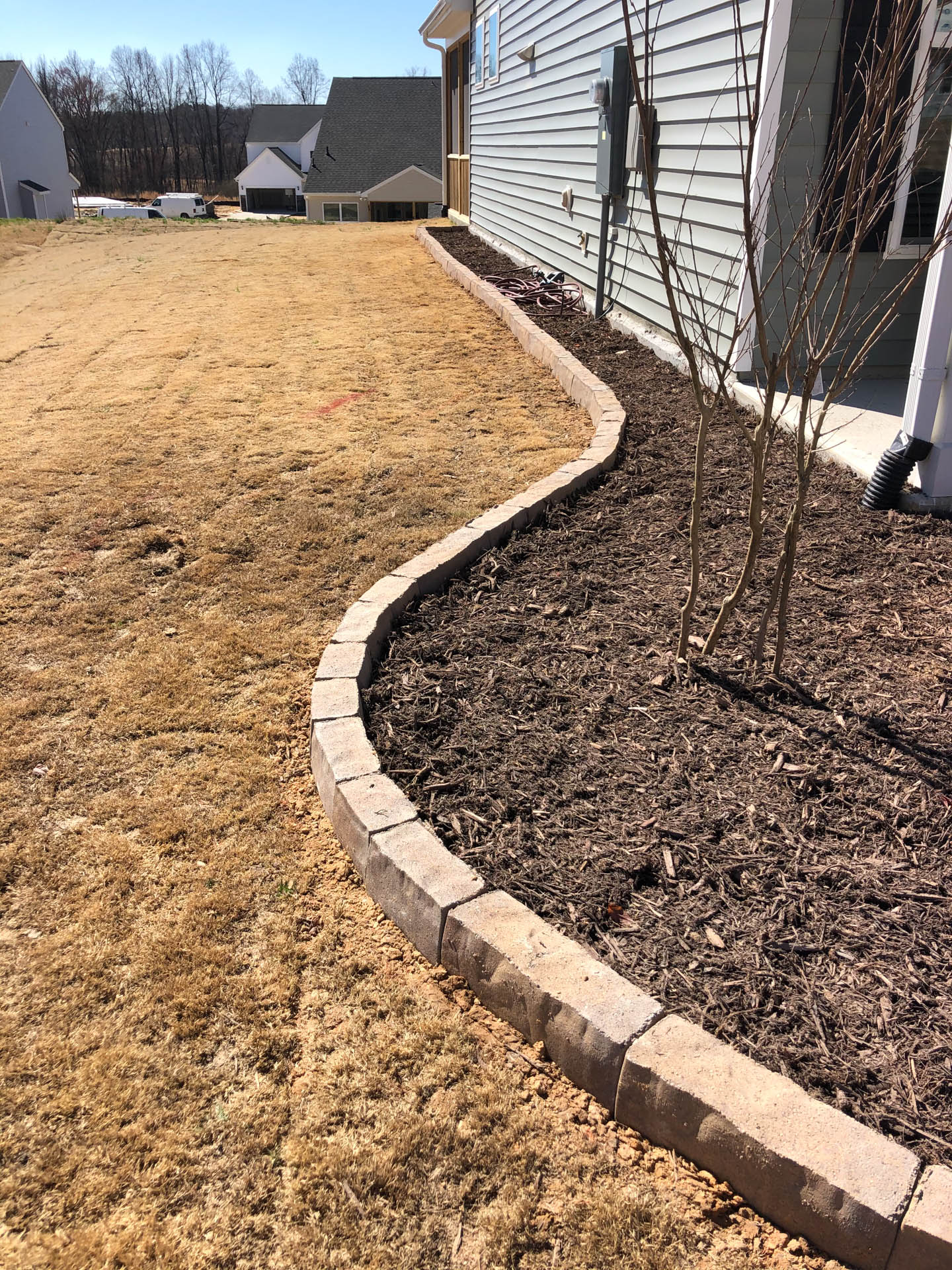 A wavy stone border separates dry grass from a mulch-covered garden bed beside a light gray house.