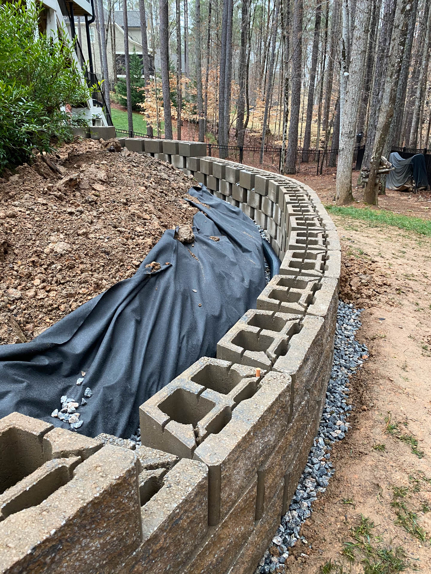 Curved retaining wall under construction. Grey blocks, black fabric, dirt, and gravel. Forest background.