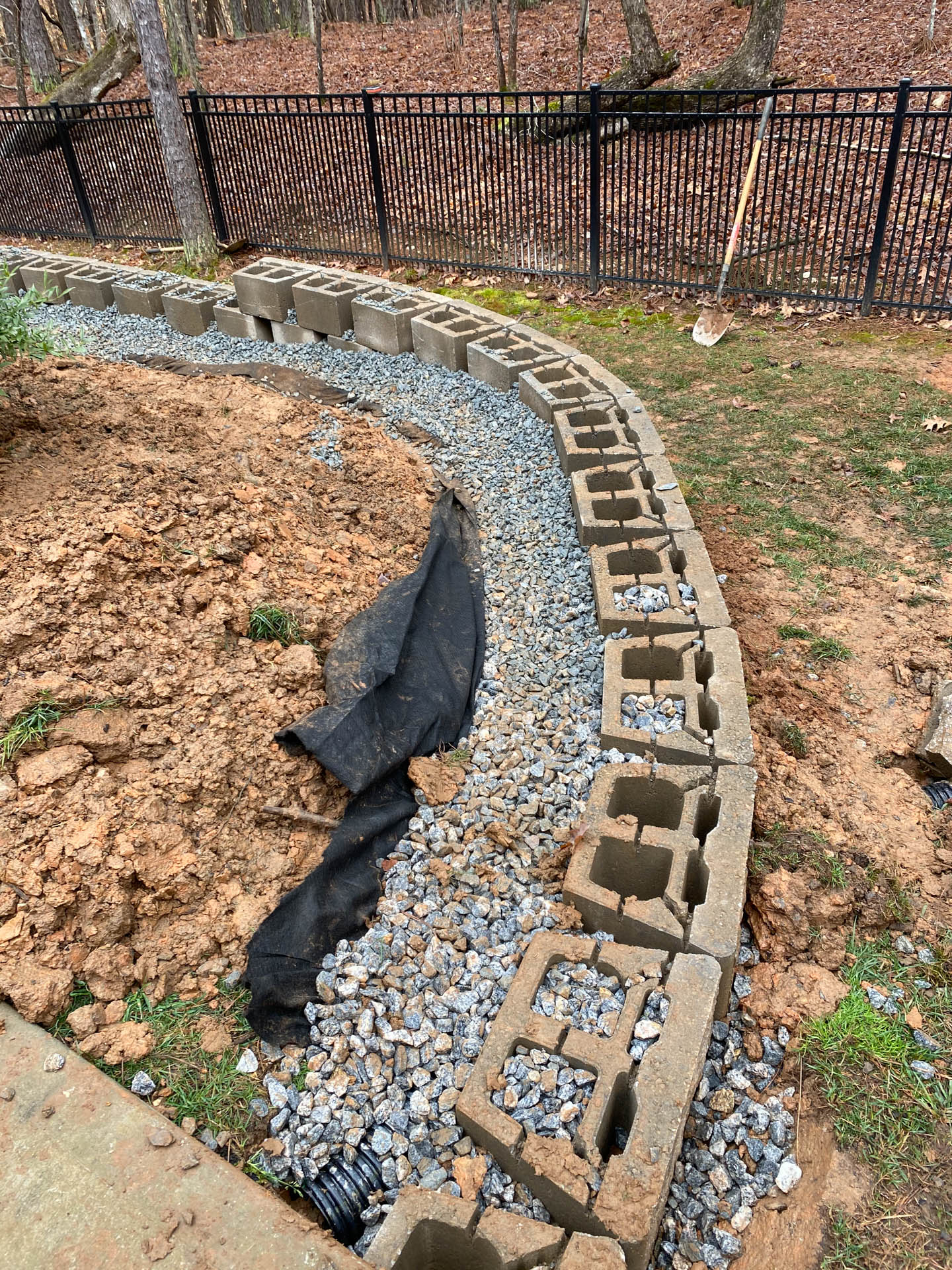Curved cinder block retaining wall under construction with gravel and fabric in yard near fence.
