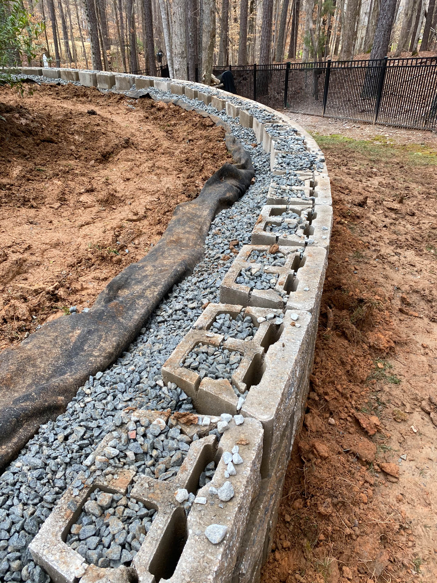 Curved retaining wall under construction with concrete blocks, gravel, and dirt.