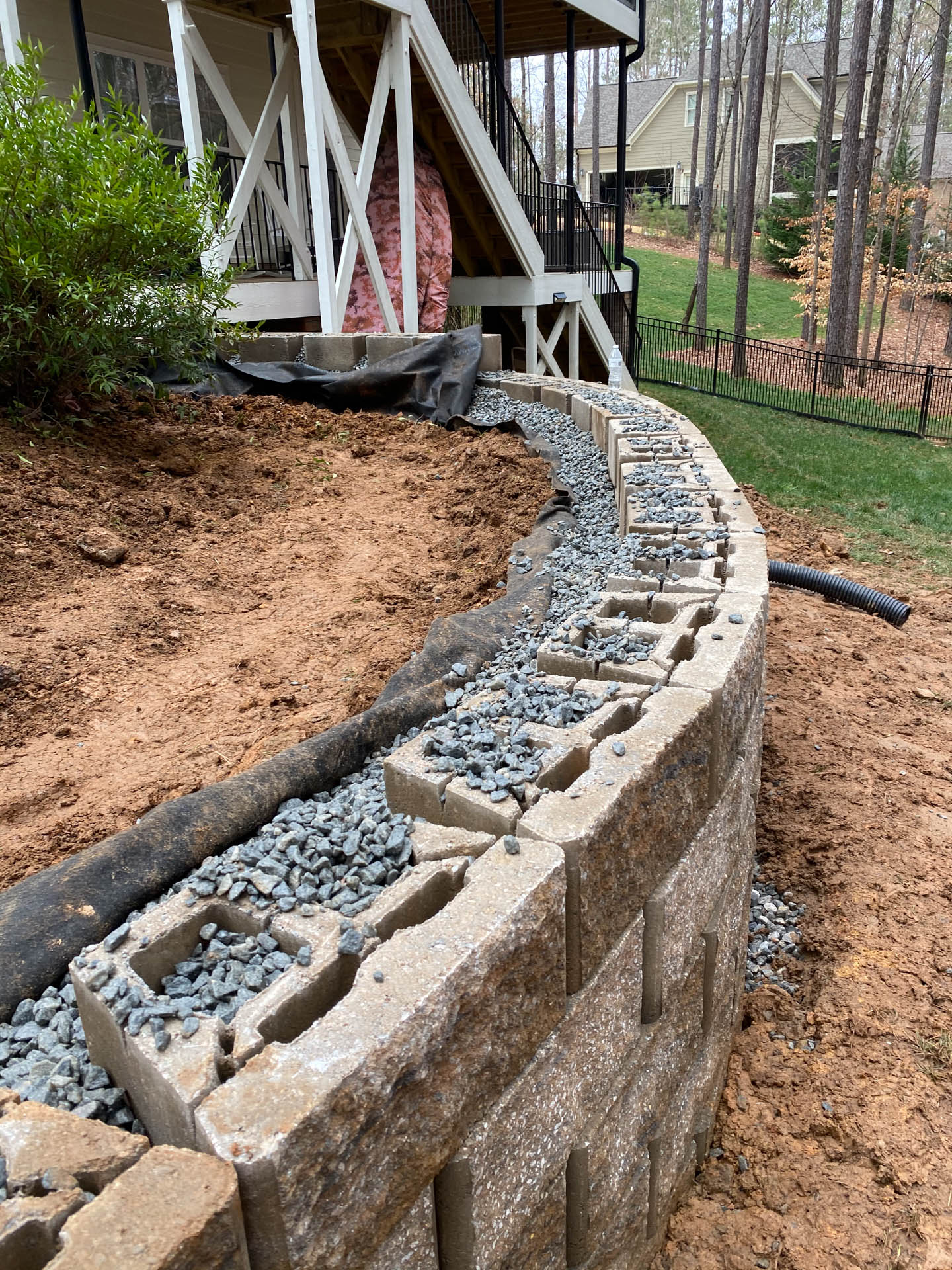 Retaining wall under construction with cinder blocks, gravel, and black fabric, next to a house and grassy area.