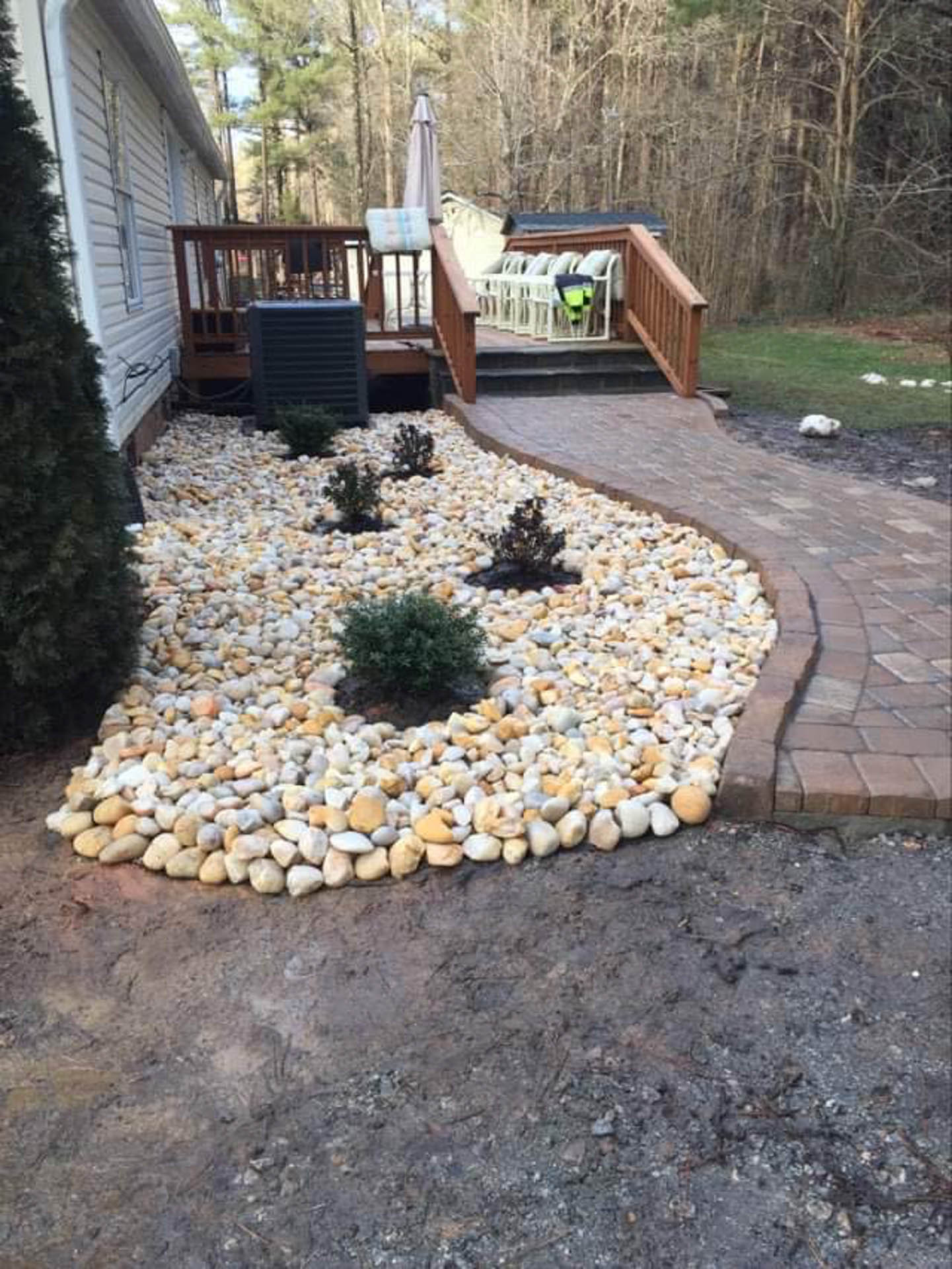 Stone-filled garden bed with small shrubs, adjacent to a brick pathway and wooden deck.