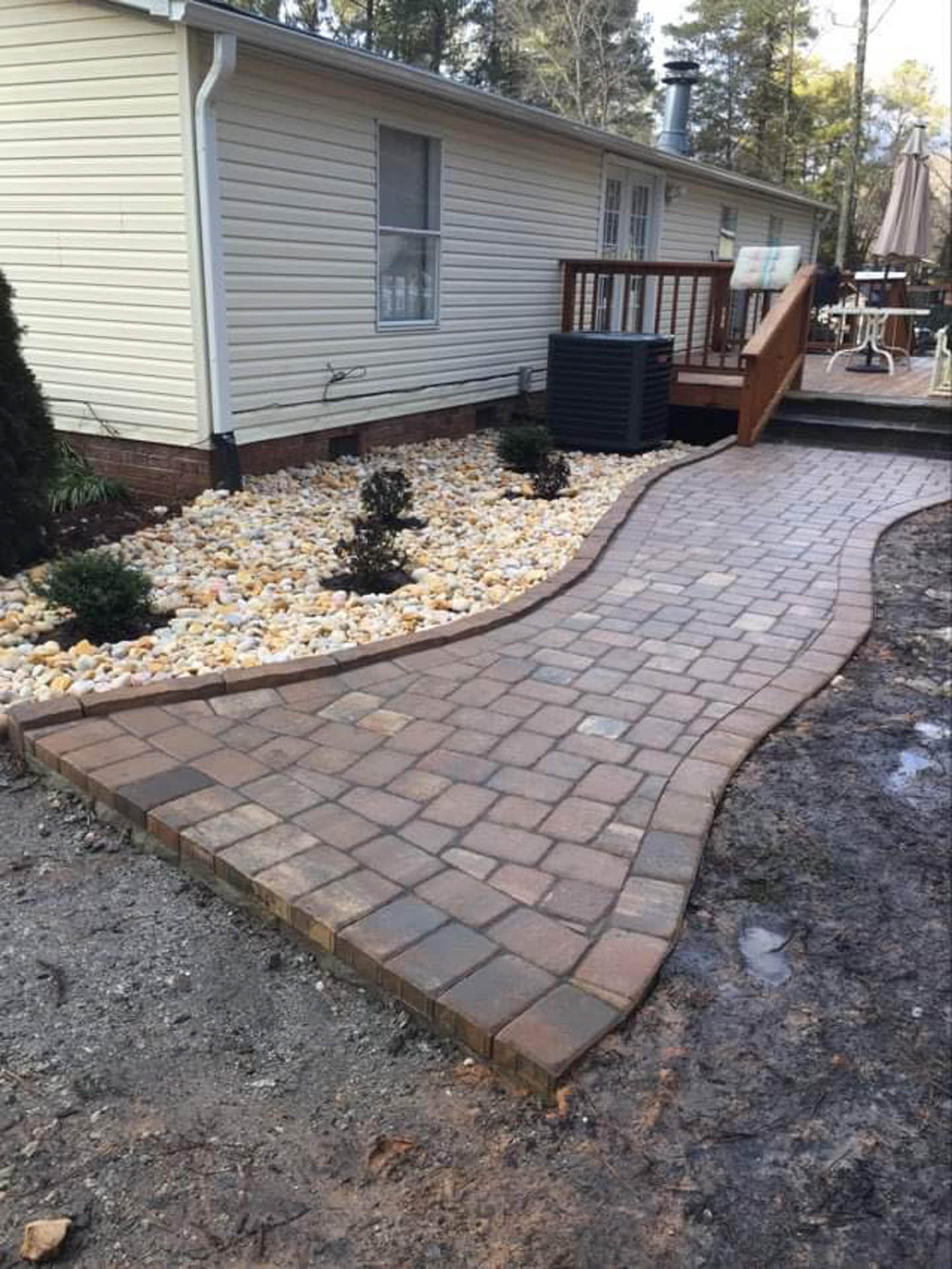 Brick pathway curves through a yard towards a deck, next to a house with landscaping.