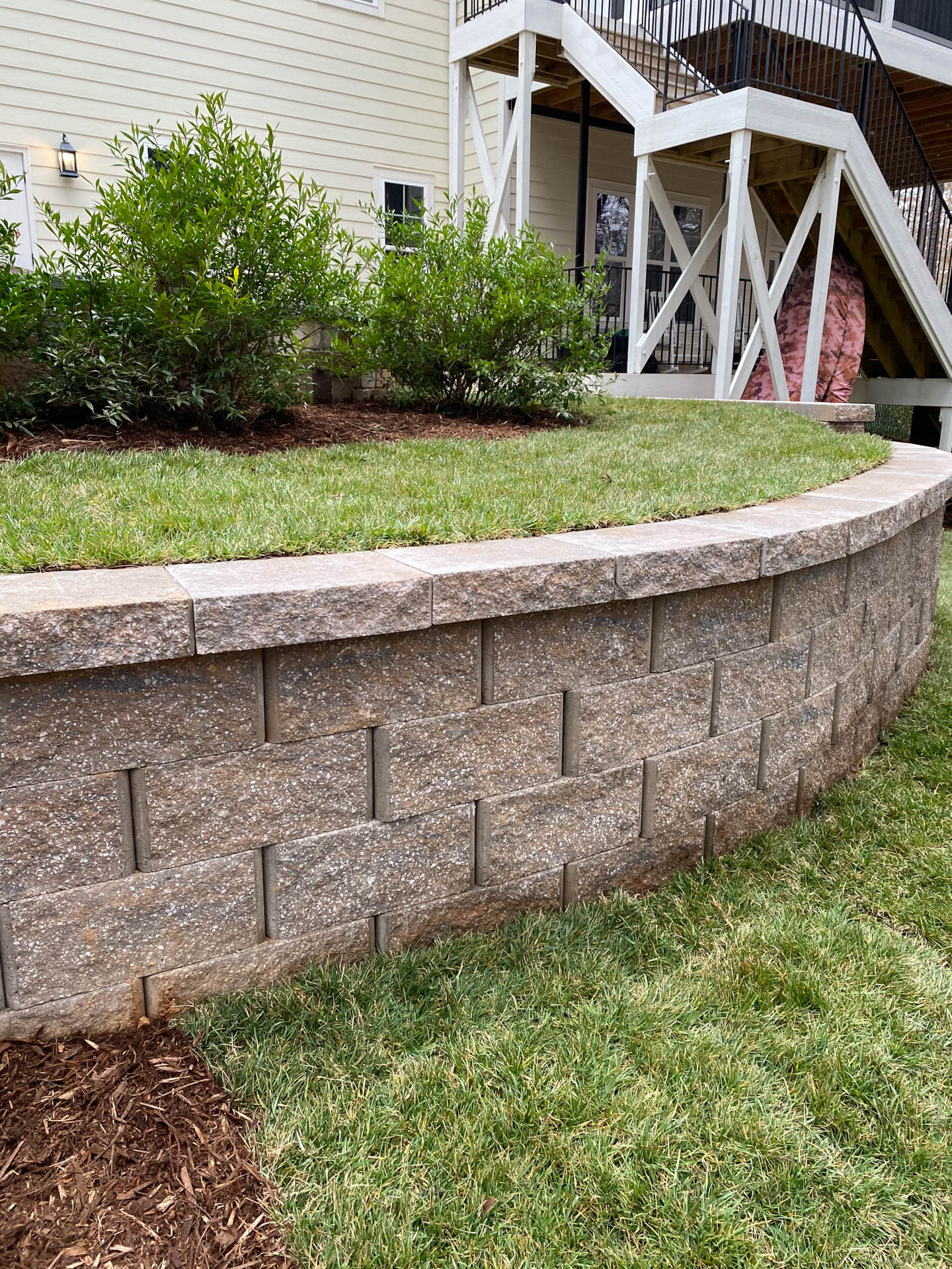 Curved retaining wall with grass on top and surrounding it, plants in the bed, stairs in the background.