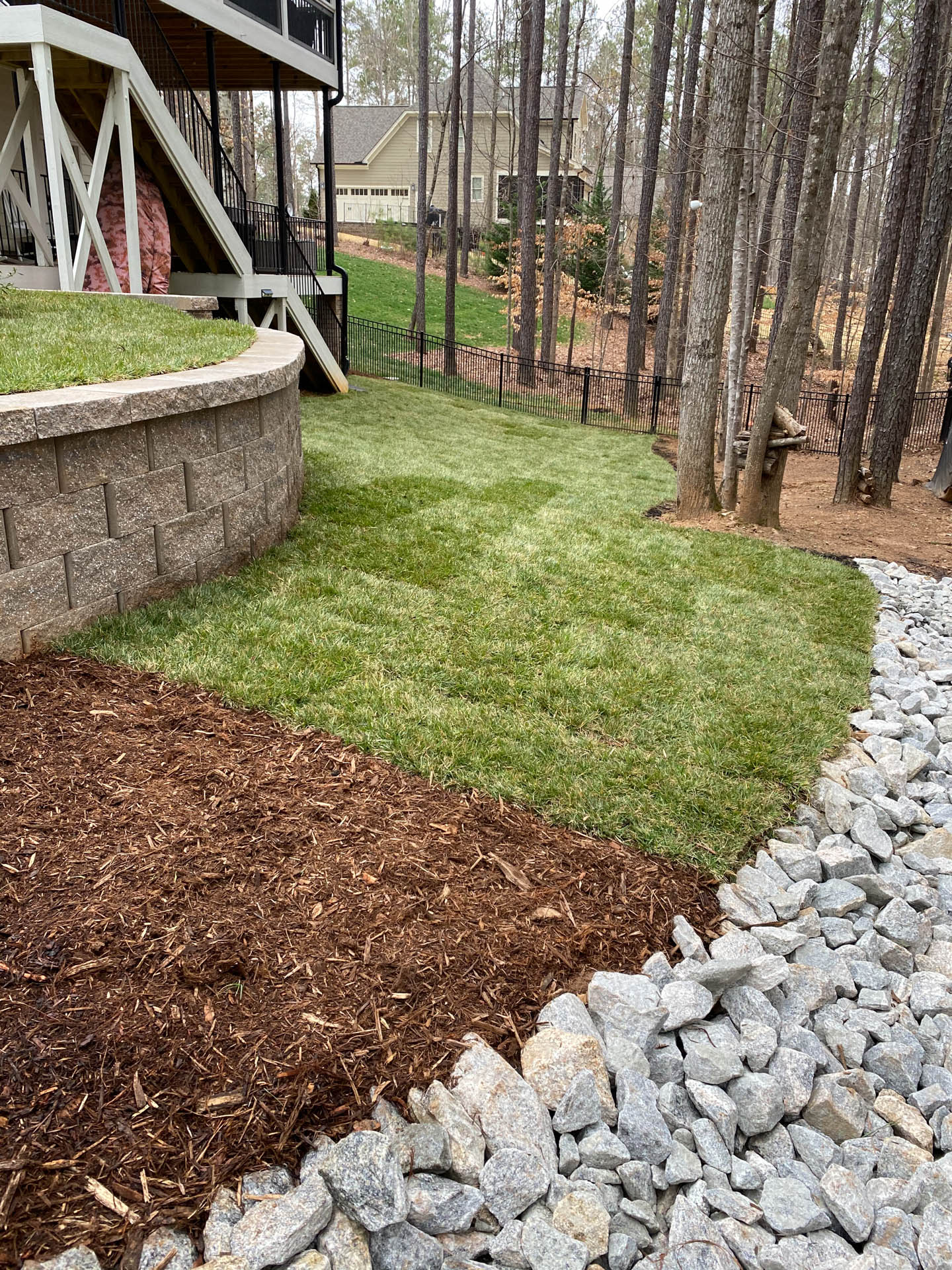 Lawn with retaining wall and mulch border transitions to a rocky stream bed in a wooded backyard setting.
