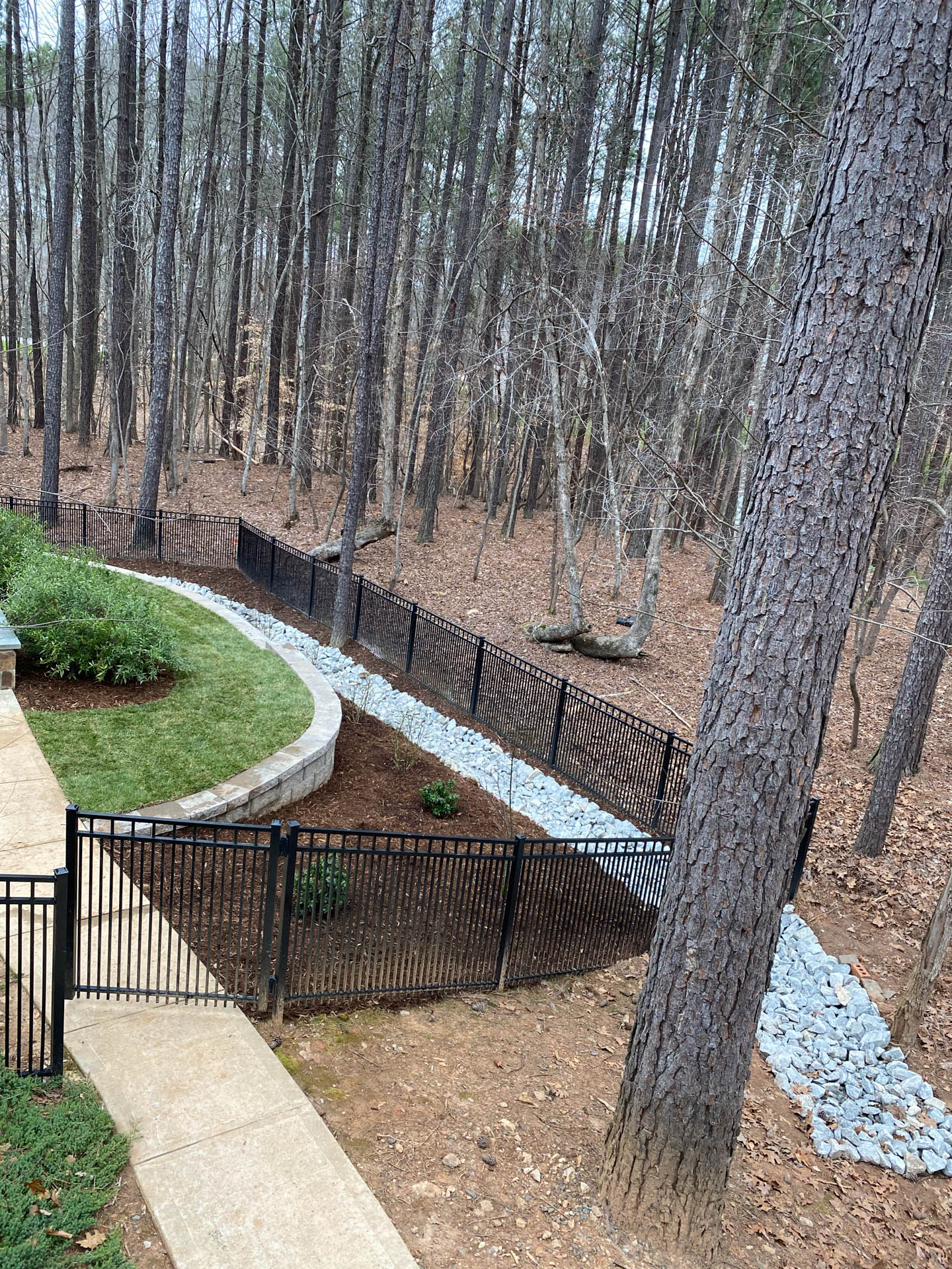 Black metal fence borders a landscaped yard, stone pathway, and a gravel drainage channel in a wooded area.