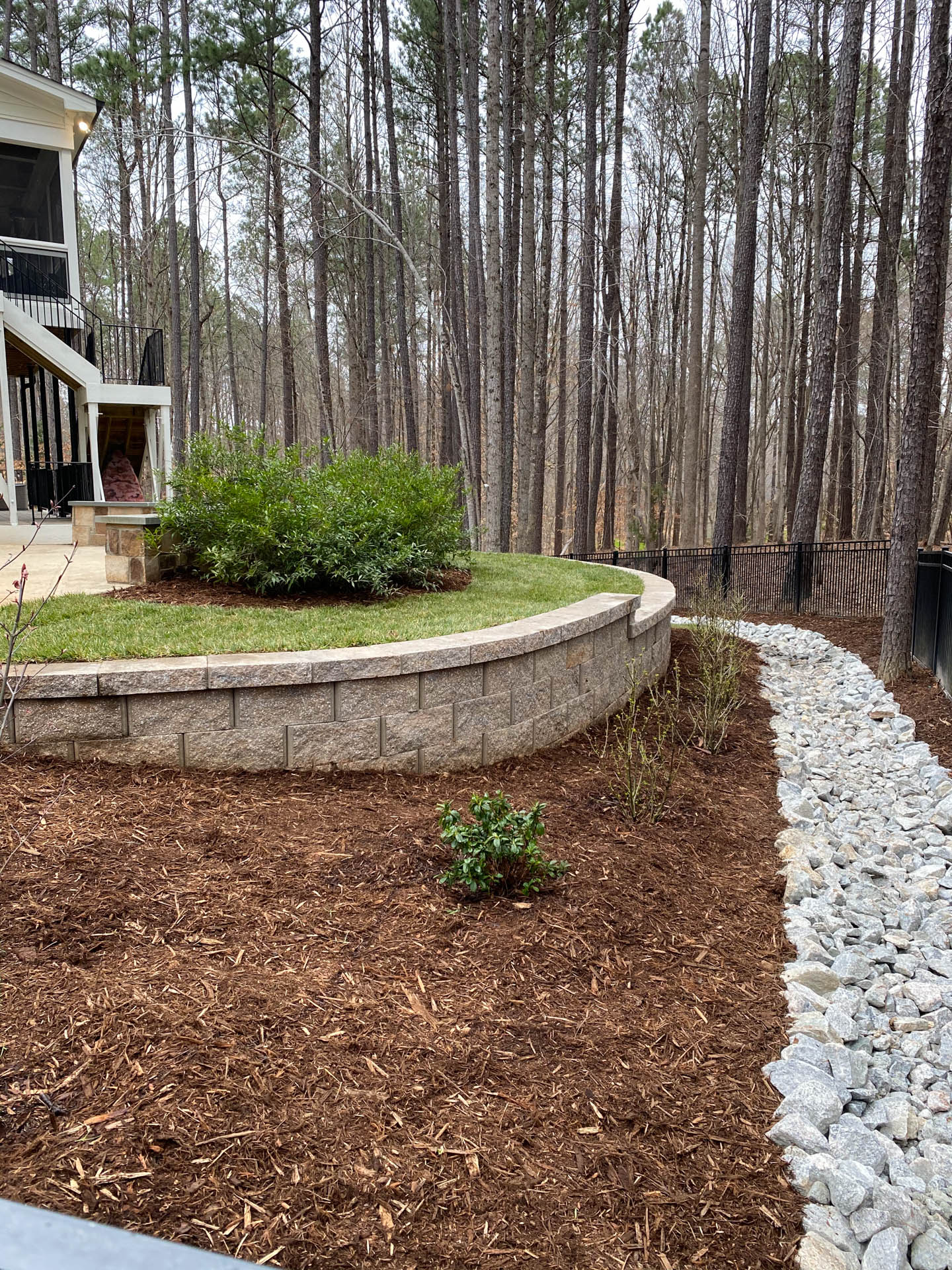Backyard with a retaining wall, mulch, shrubs, and a gravel path leading into a wooded area.