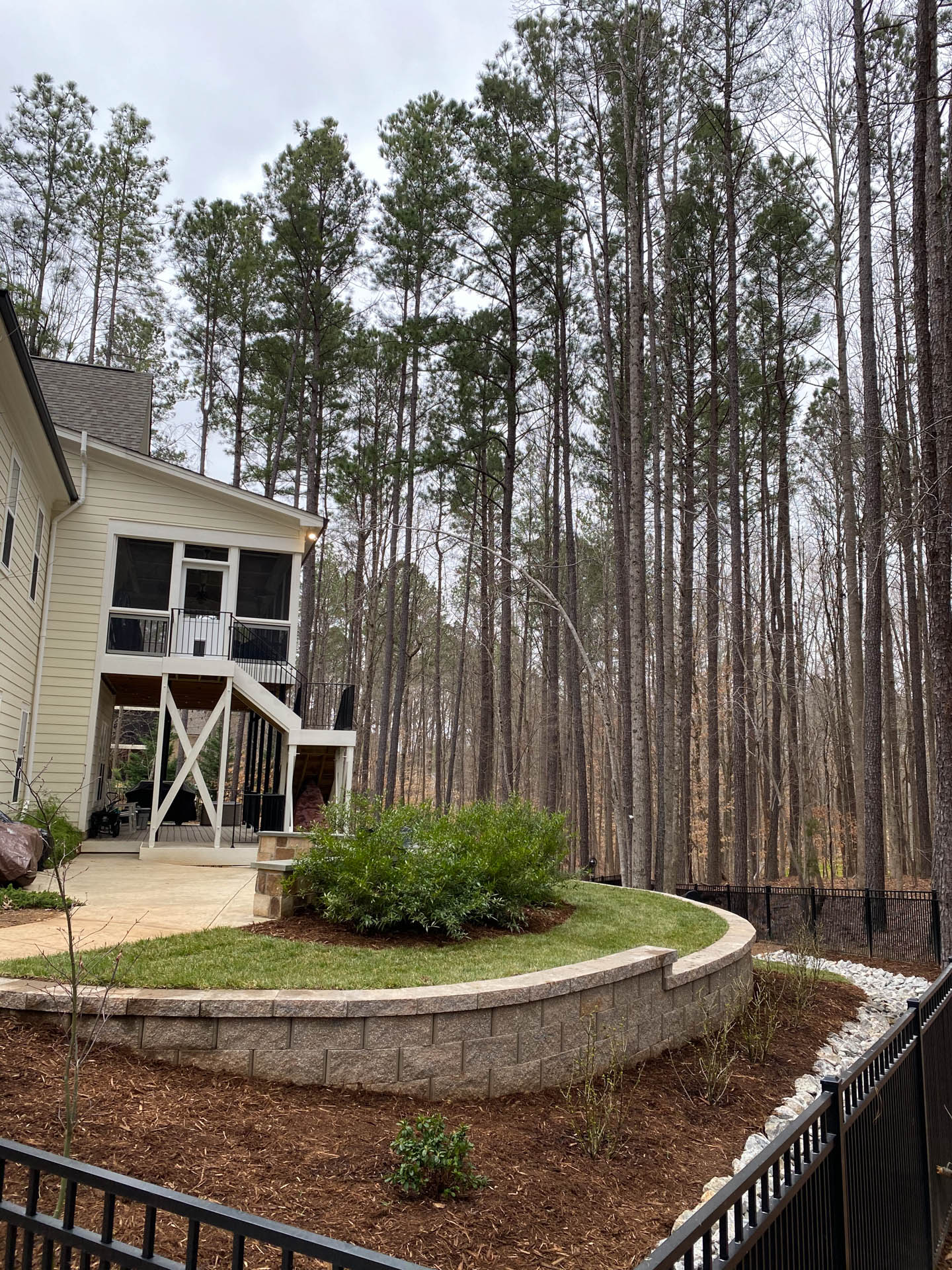 Backyard with a retaining wall, a porch, trees, and a black fence.
