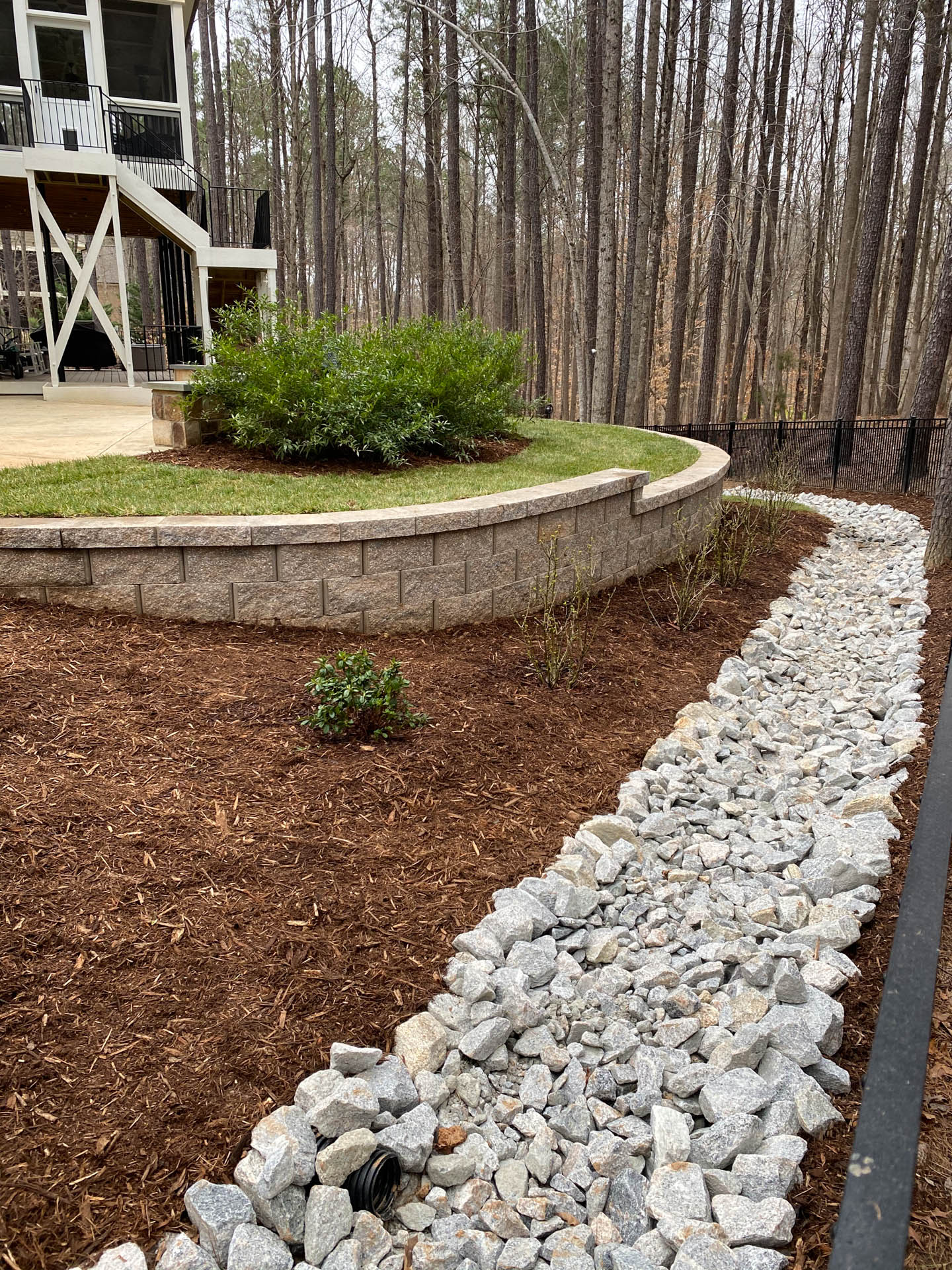 Stone-lined drainage channel alongside a landscaped area with retaining wall, mulch, and greenery.