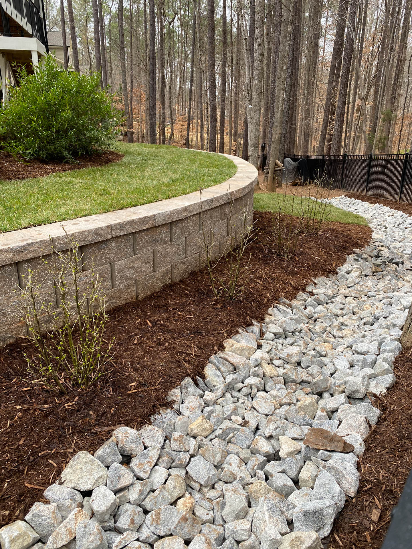 Stone-lined drainage channel with rock, mulch, and retaining wall next to grassy yard and trees.