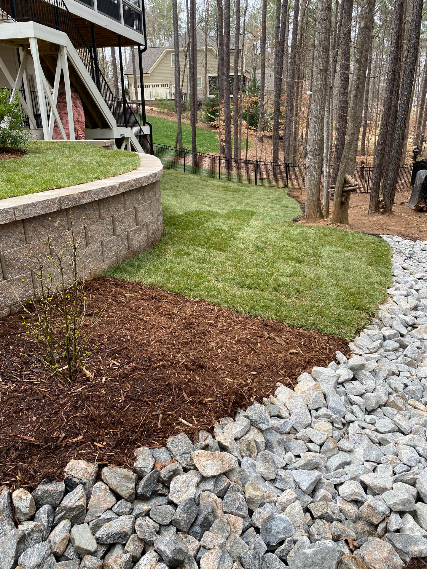 A landscaped yard with a retaining wall, mulch, and stone path next to a lawn and trees.